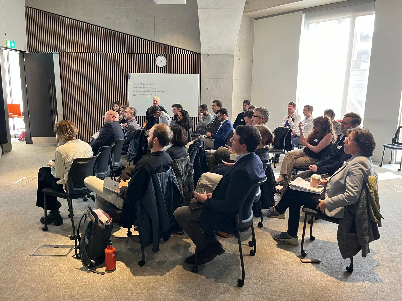 carbon at risk A group of people attending a conference or seminar in a bright room with large windows, sitting on black chairs facing forward, some taking notes and holding drinks, with a whiteboard and clock on the wall behind them.