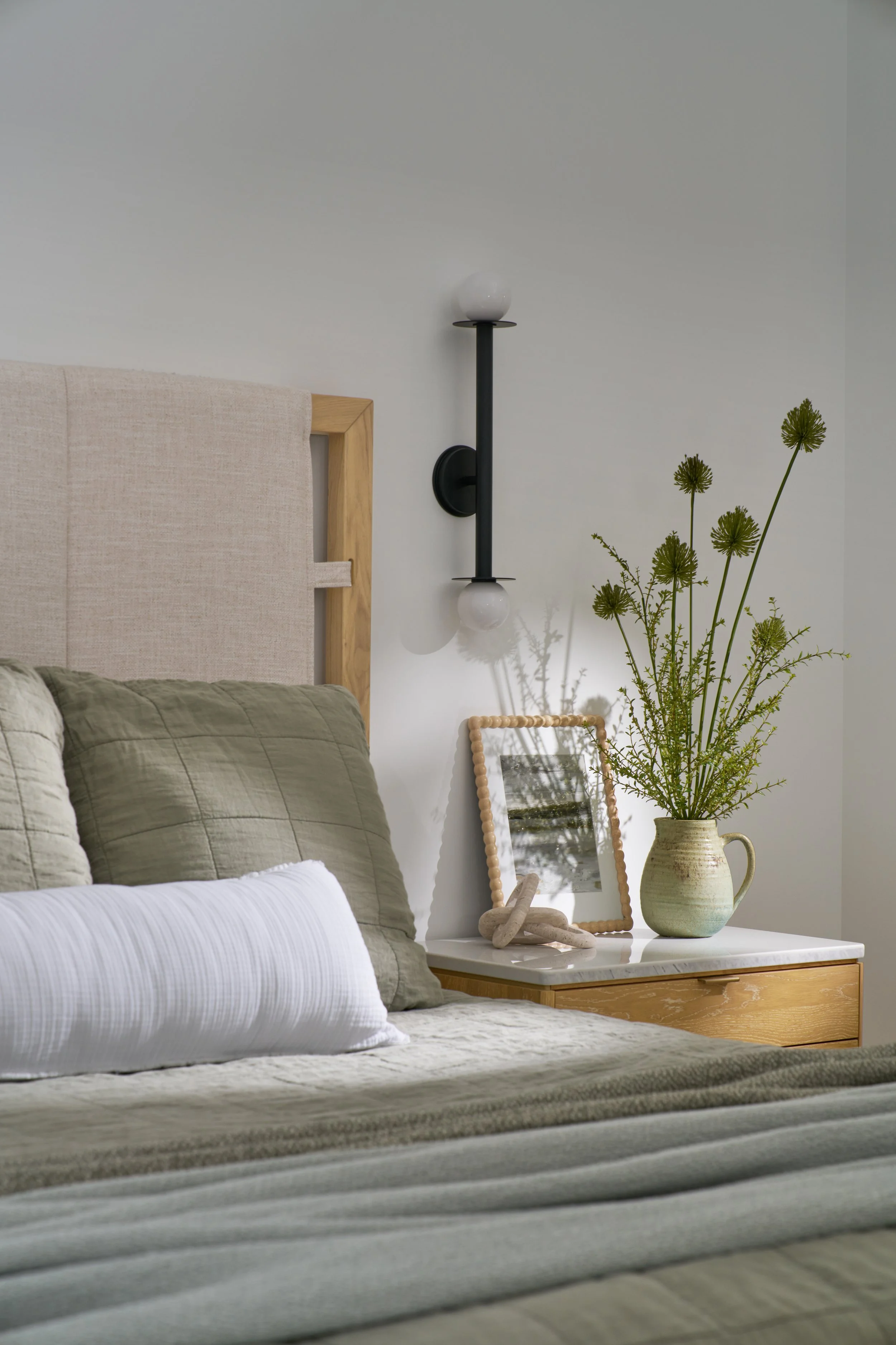 Natural modern bedroom vignette with upholstered headboard, wood and marble nightstand, and geometric wall sconce