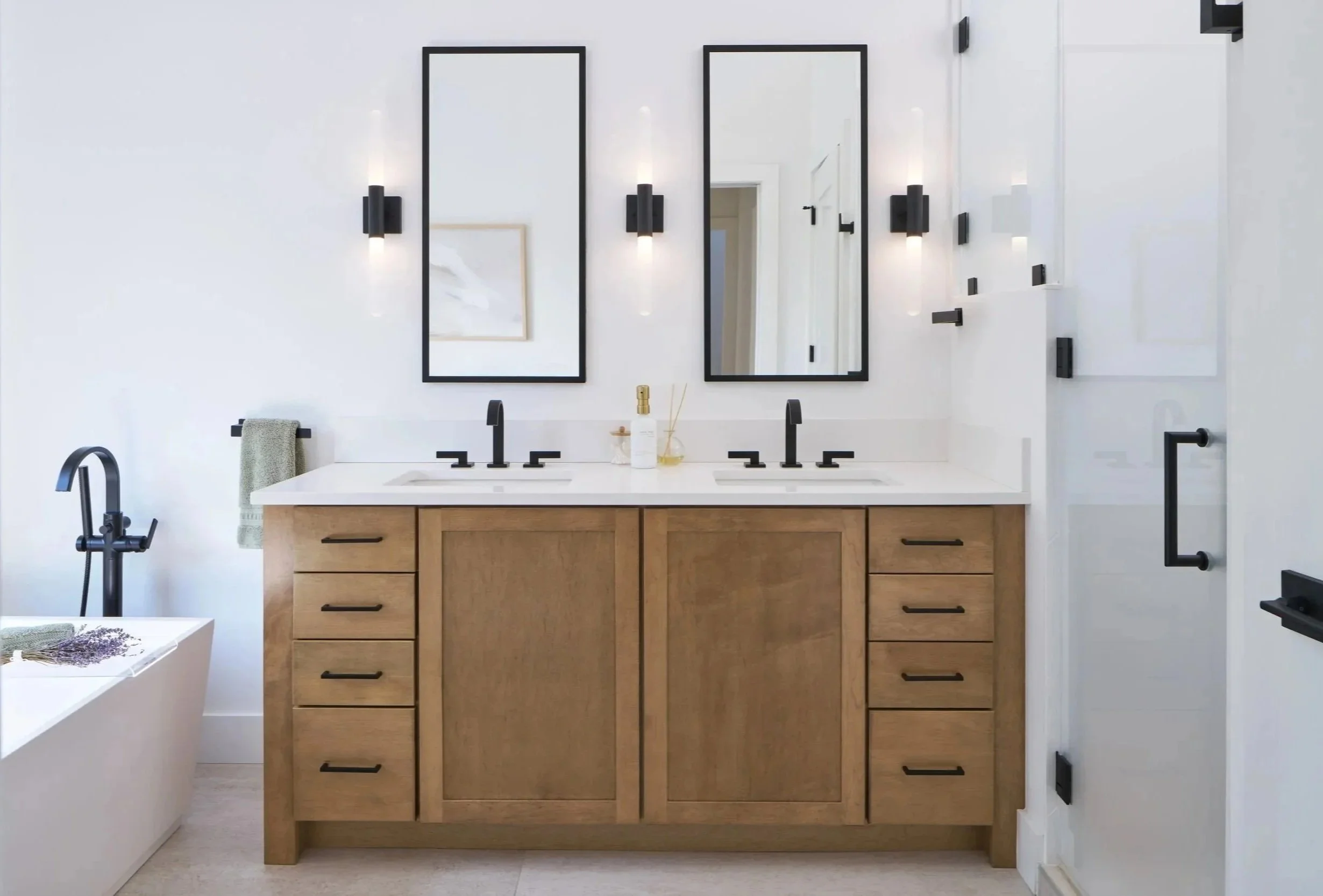 Bright modern bathroom with natural wood double vanity, quartz countertop, matte black plumbing fixtures, wall sconces, large format tile floor, and frosted shower and soaker tub