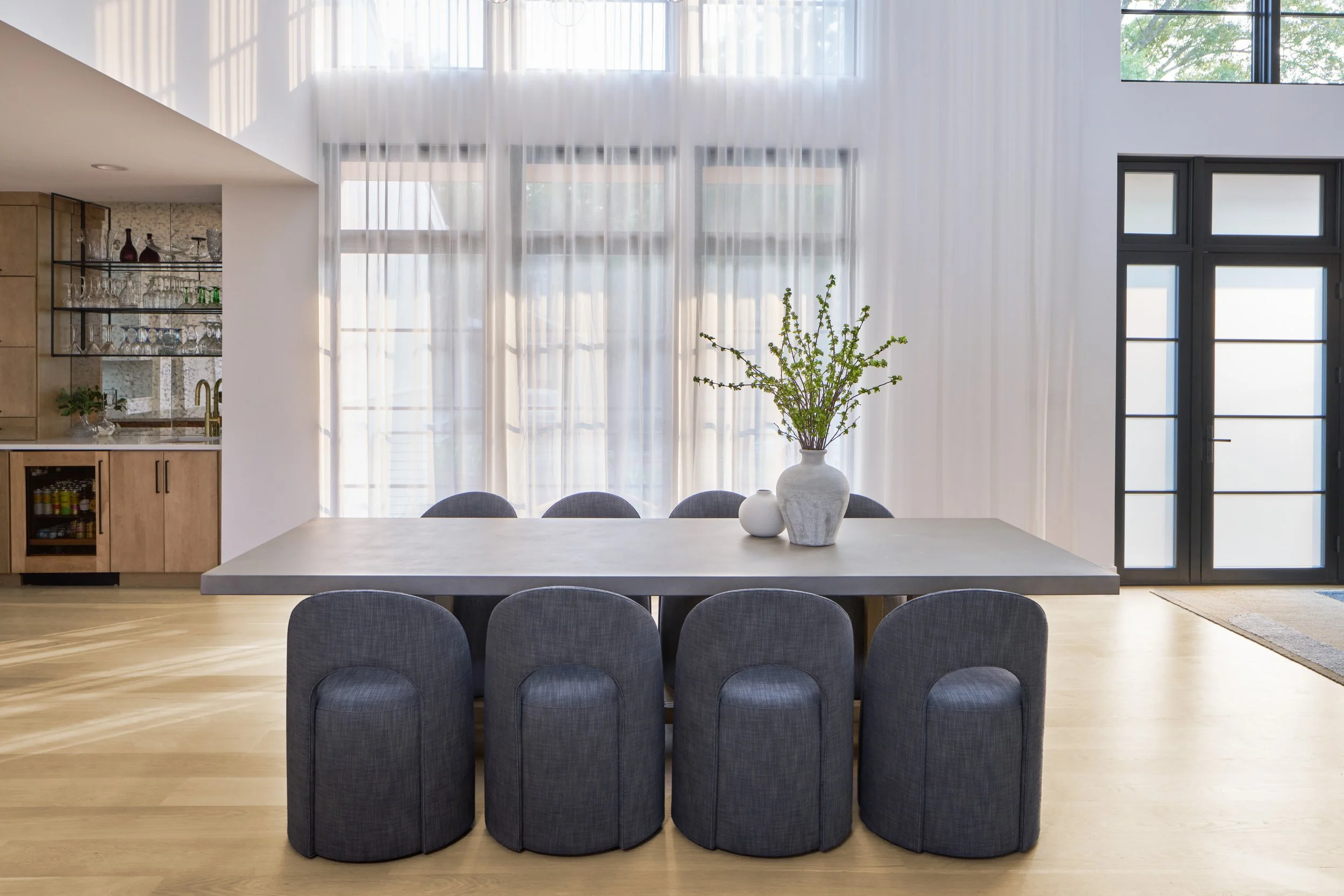 Modern dining room with concrete and oak table, geometric upholstered chairs, and soft diffusing curtains over floor to ceiling windows, featuring a wet bar