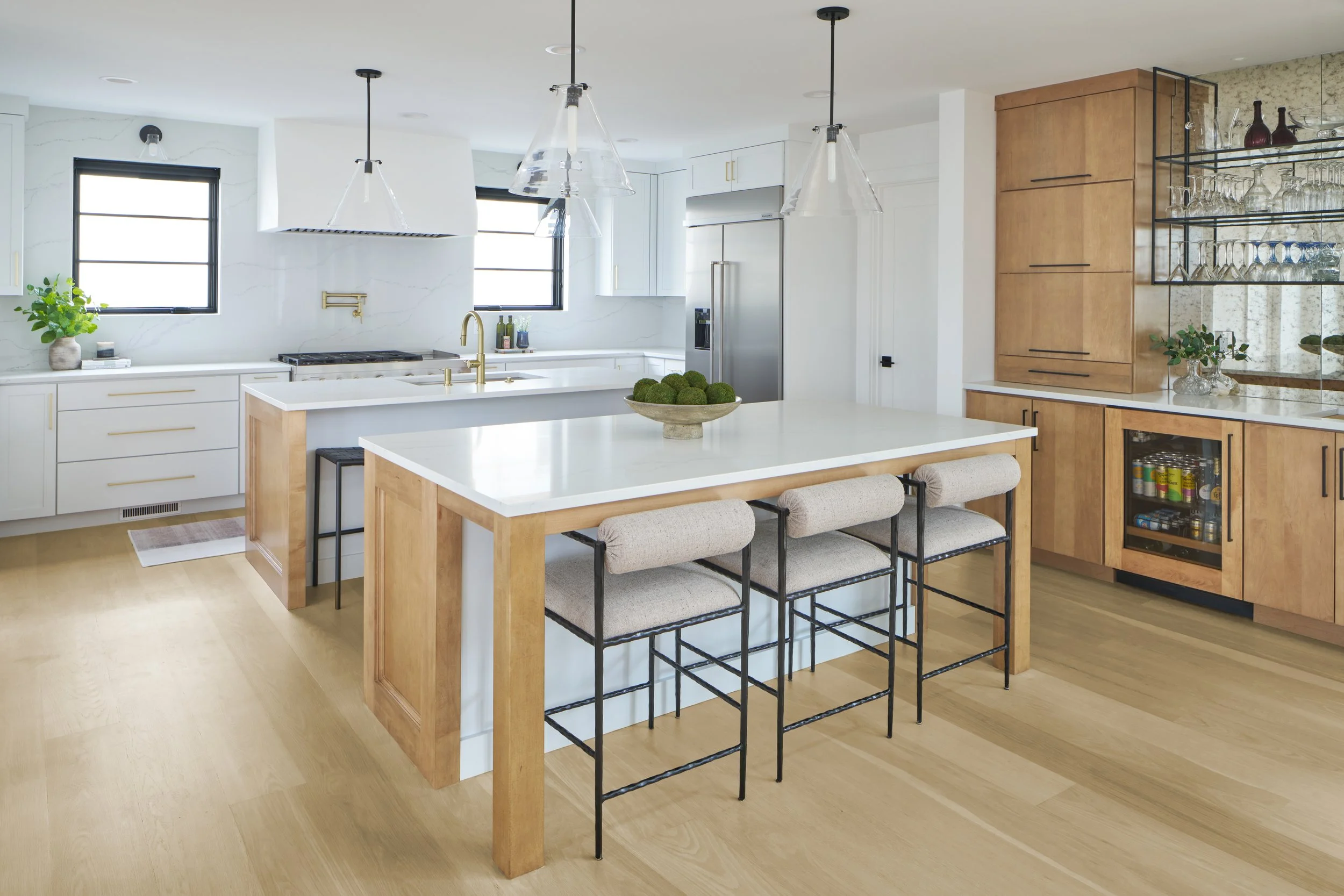Bright contemporary kitchen with double islands, mixed white and natural wood cabinetry, wide white oak flooring, marble/quartz counter, and an antiqued mirrored modern wet bar