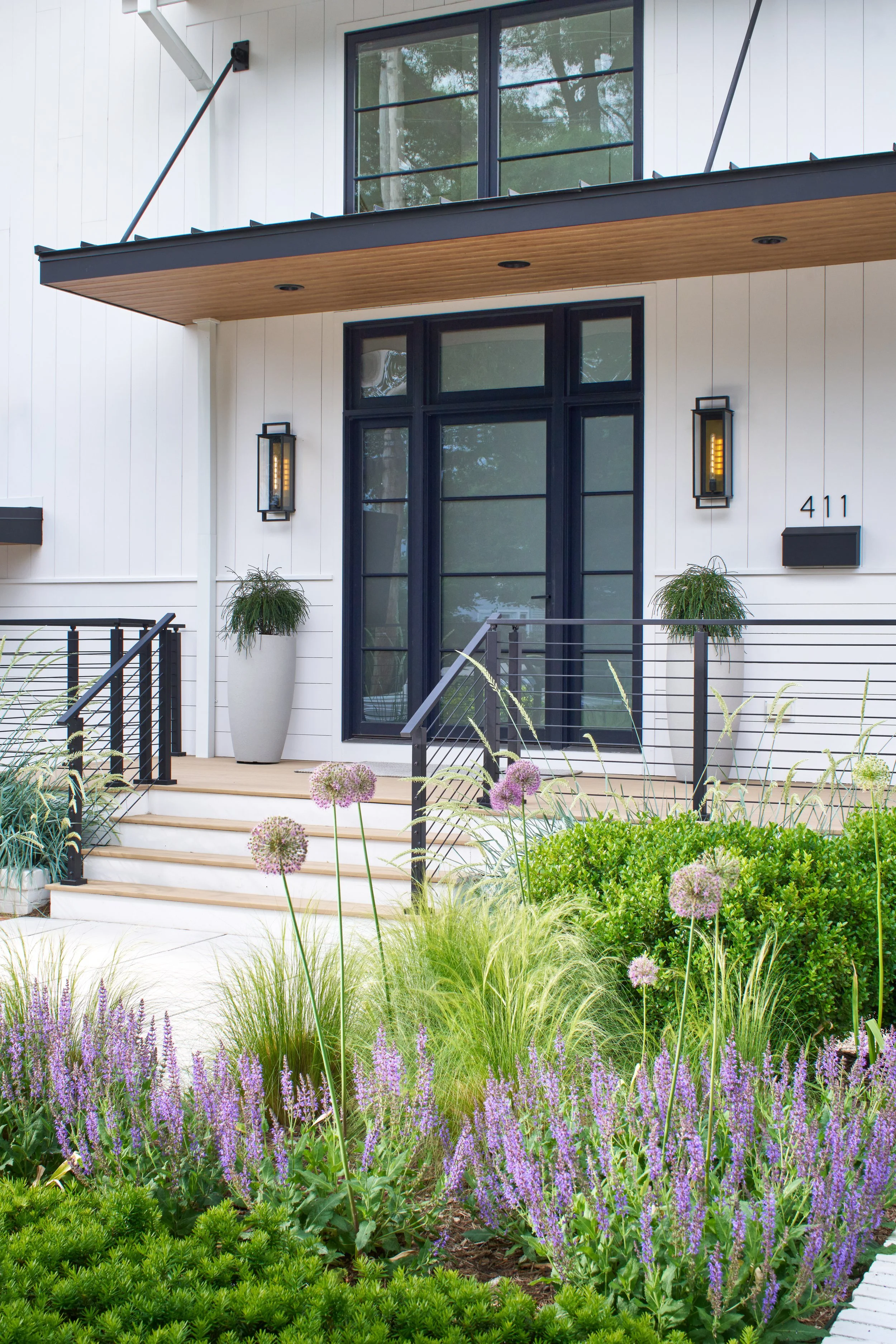 White modern transitional home exterior with natural wood tones, black iron doors, modern glass brass lanterns, and coastal landscaped entry