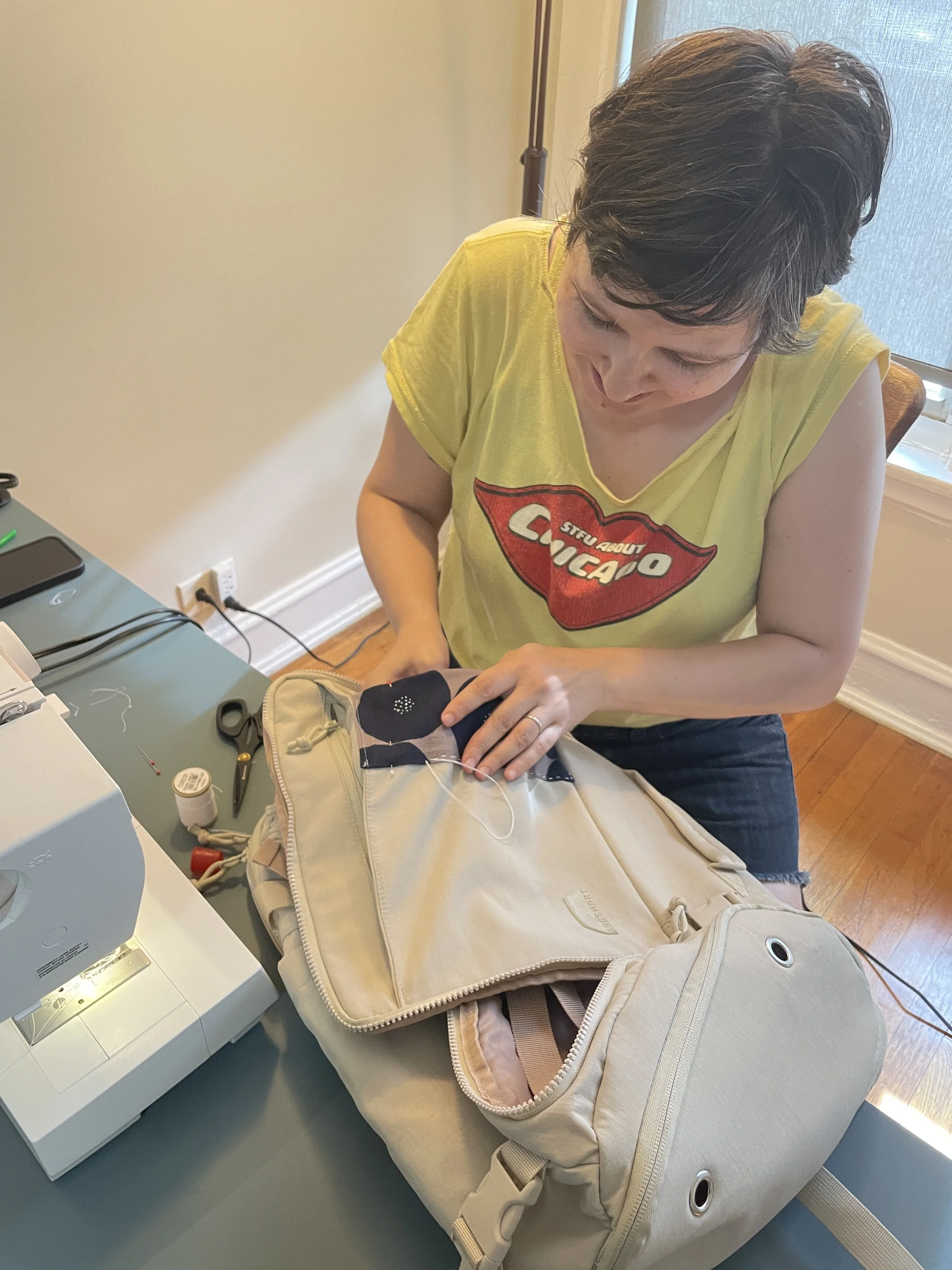 Woman sewing a patch onto a beige backpack at a green table.