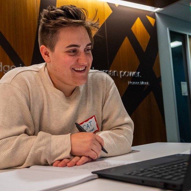 Consultant with short hair wearing a beige sweater, smiling during a conversation or meeting, sitting at a table with a laptop and papers in front of them.