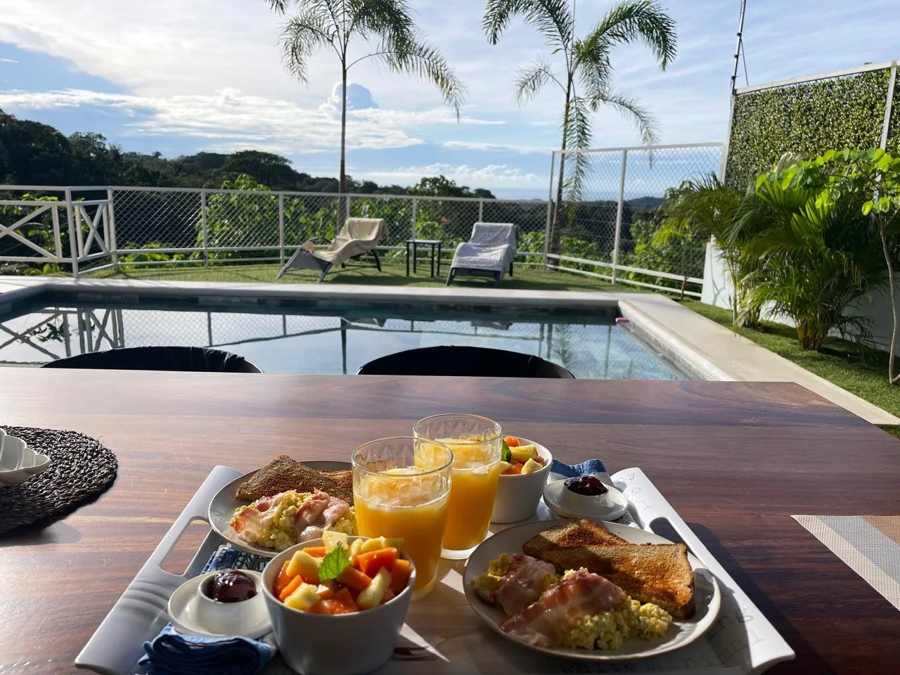 Petit déjeuner sur une table en bois avec vue sur une piscine, des chaises longues et un paysage verdoyant sous un ciel partiellement nuageux.