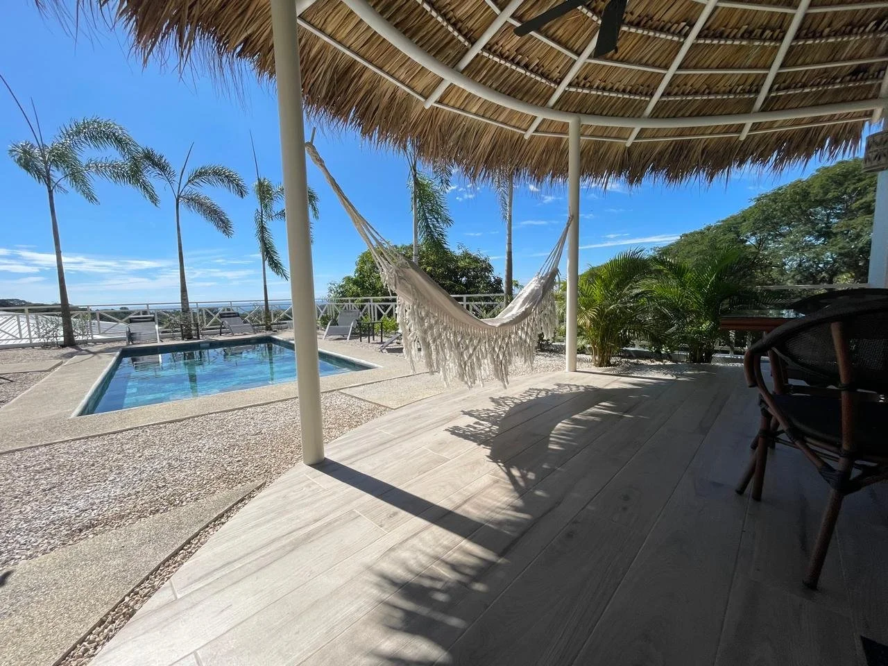 Terrasse sous un toit de chaume avec une balançoire en tissu blanc, une petite piscine, des chaises longues, des plantes vertes, en bord de mer sous un ciel bleu.