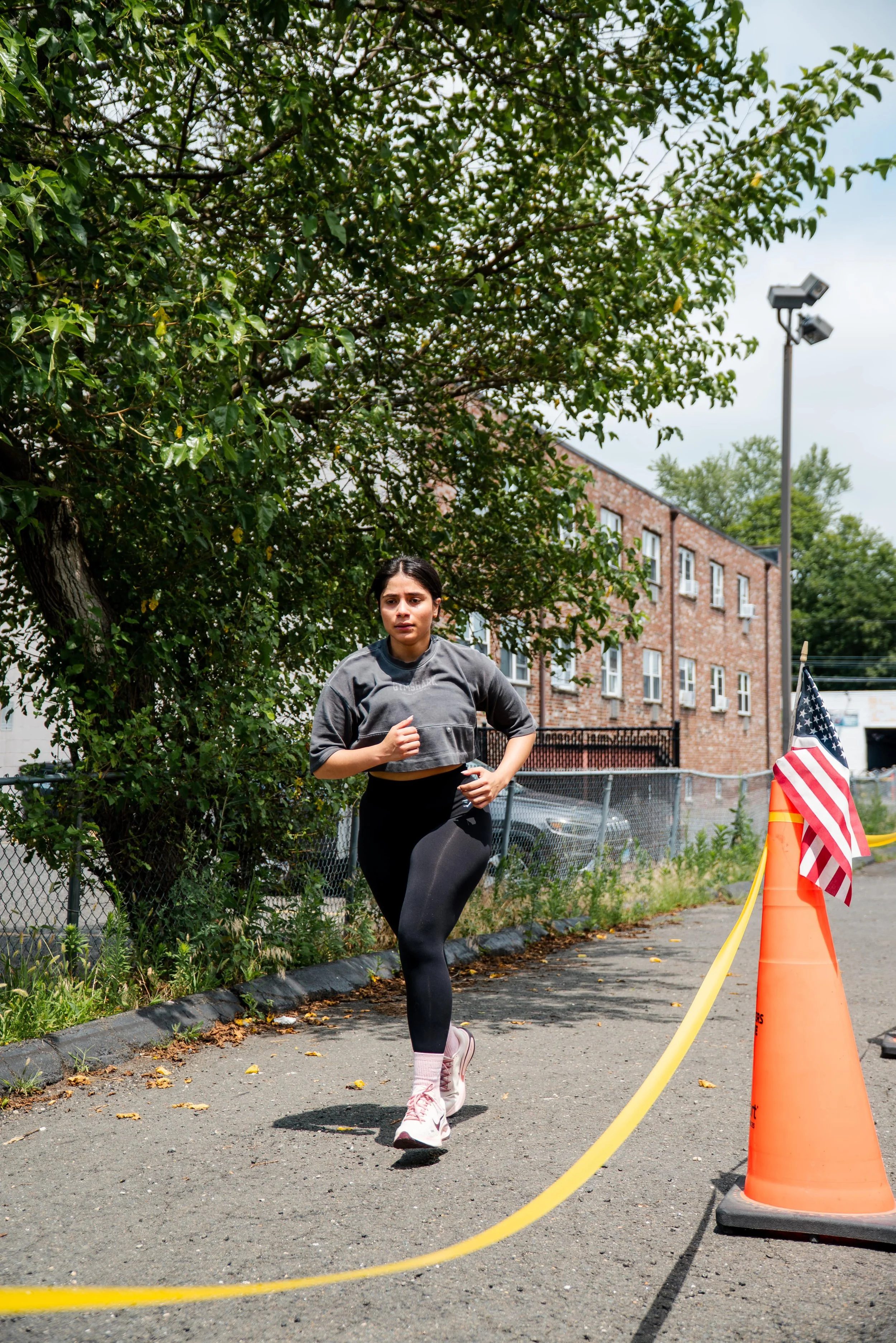 A woman running outdoors during the daytime, wearing a gray top, black leggings, pink socks, and white running shoes, on a paved path with a fence, trees, and a building in the background, next to an orange safety cone and an American flag.
