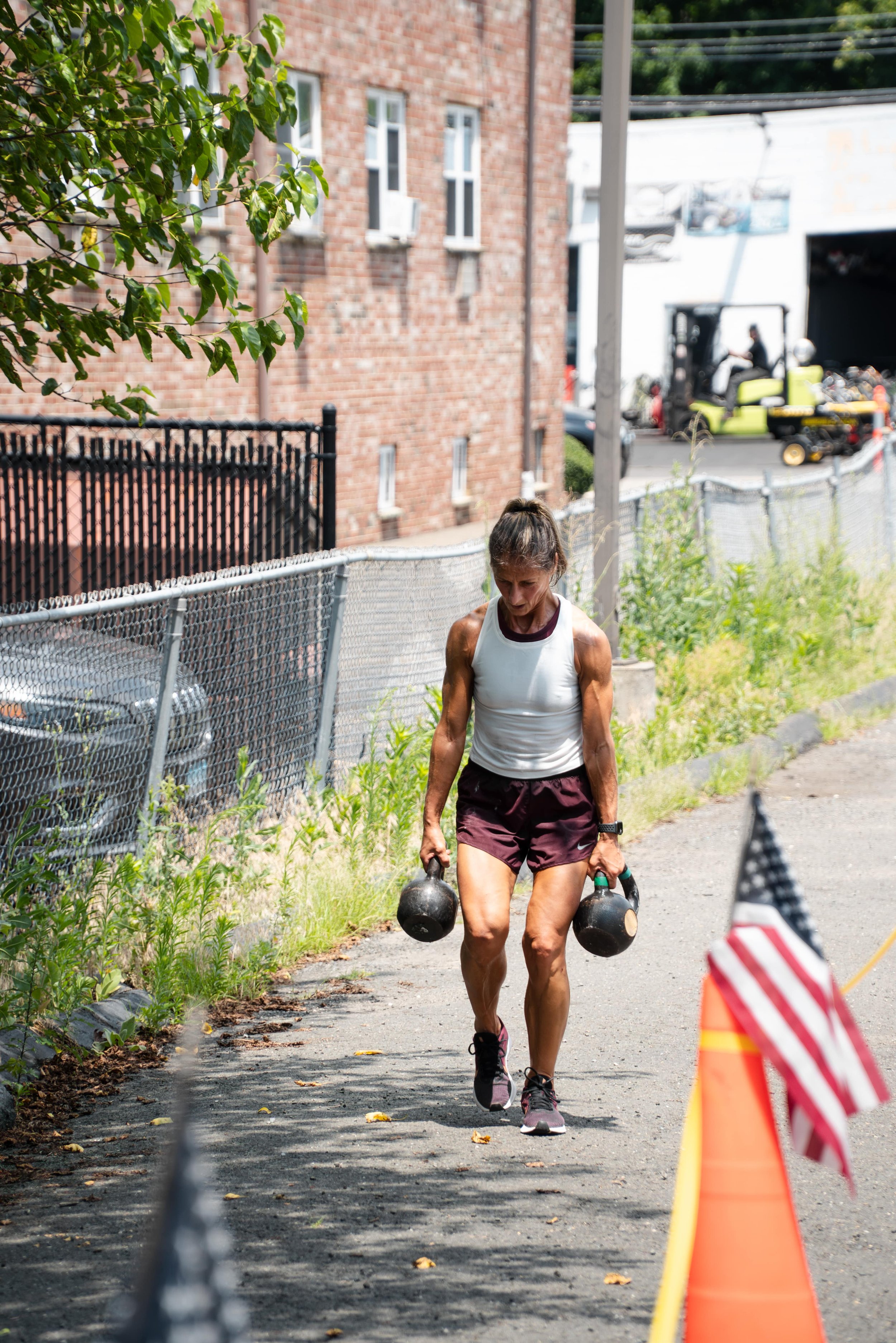 A woman in athletic attire lifting kettlebells outdoors during a workout or competition, with flags and orange cones marking the area.