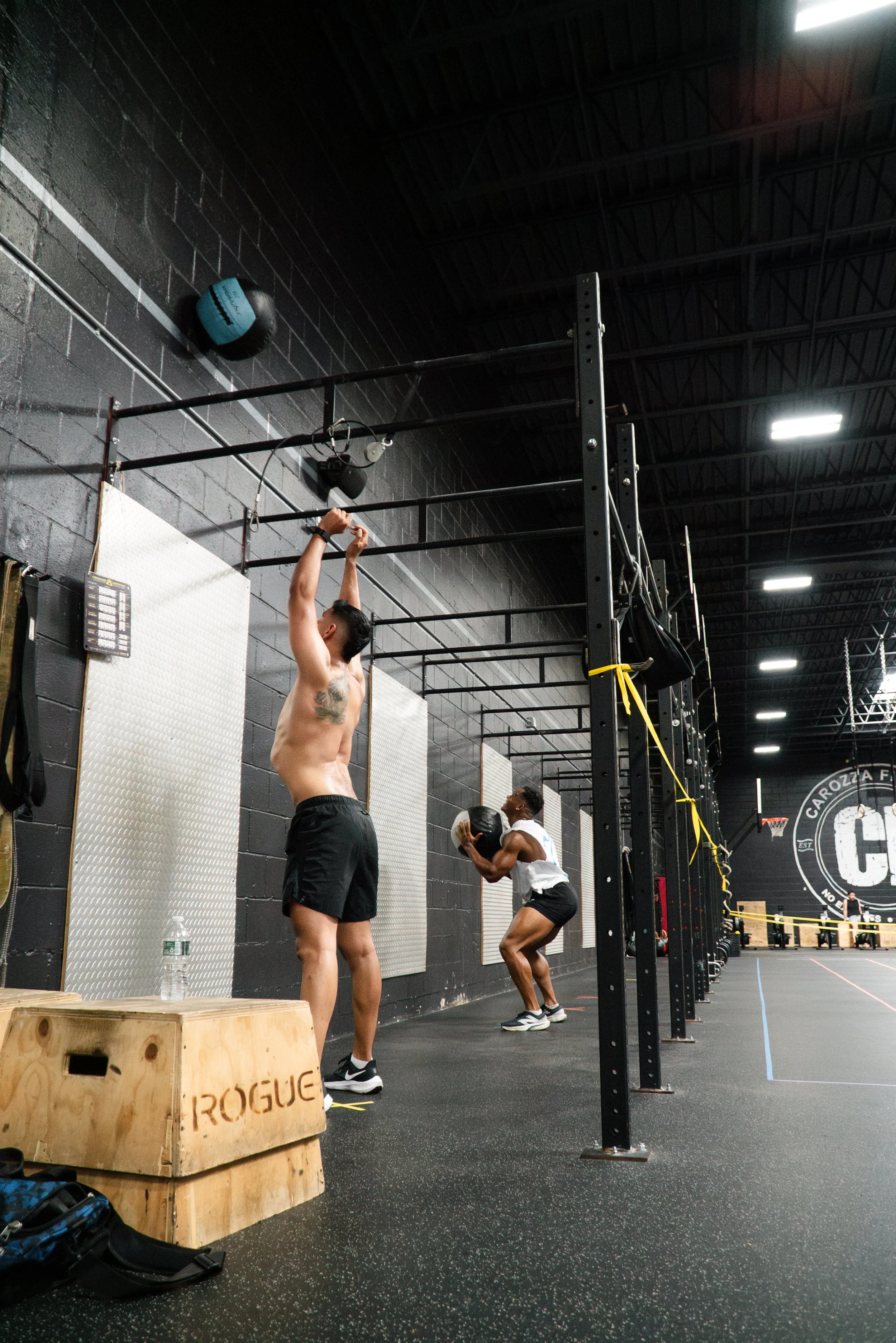 Two people working out in a gym with black walls and high ceiling, using wall bars and holding medicine balls. The person in the foreground is shirtless and stretching with arms raised, while the person in the background is squatting with a medicine