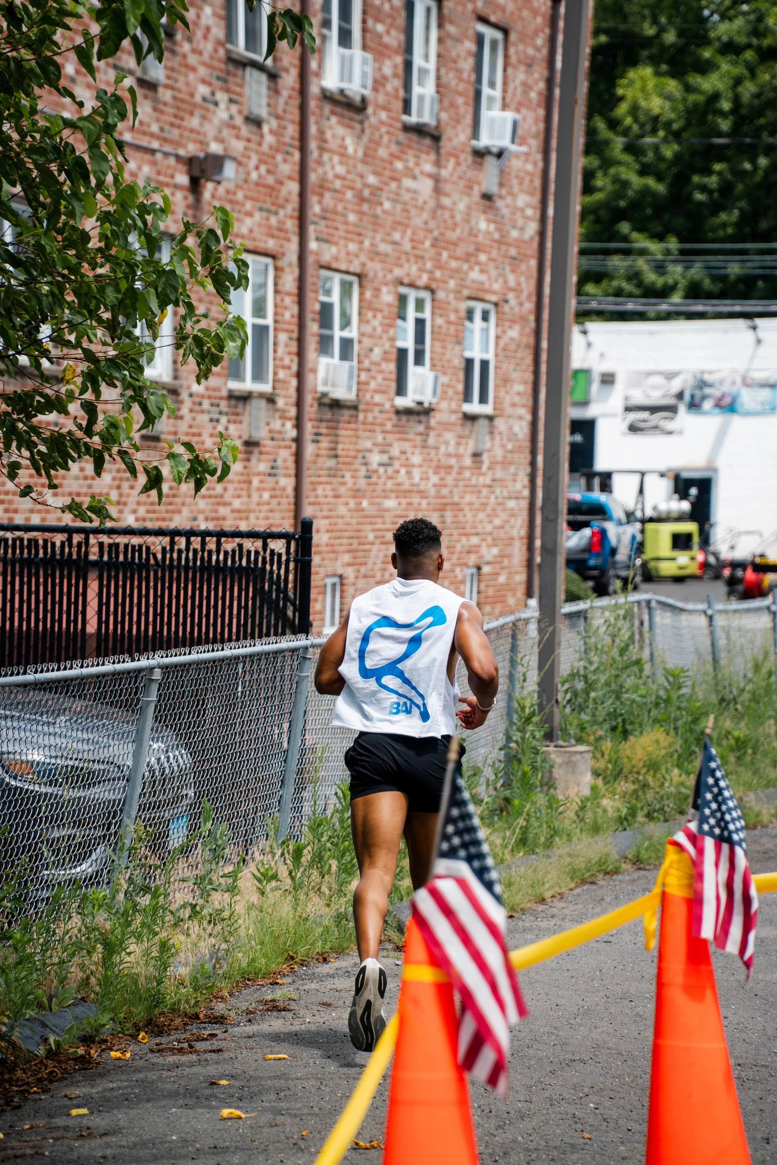 A man running outdoors along a fence with orange cones and small American flags, in front of a brick building with multiple windows and air conditioning units.