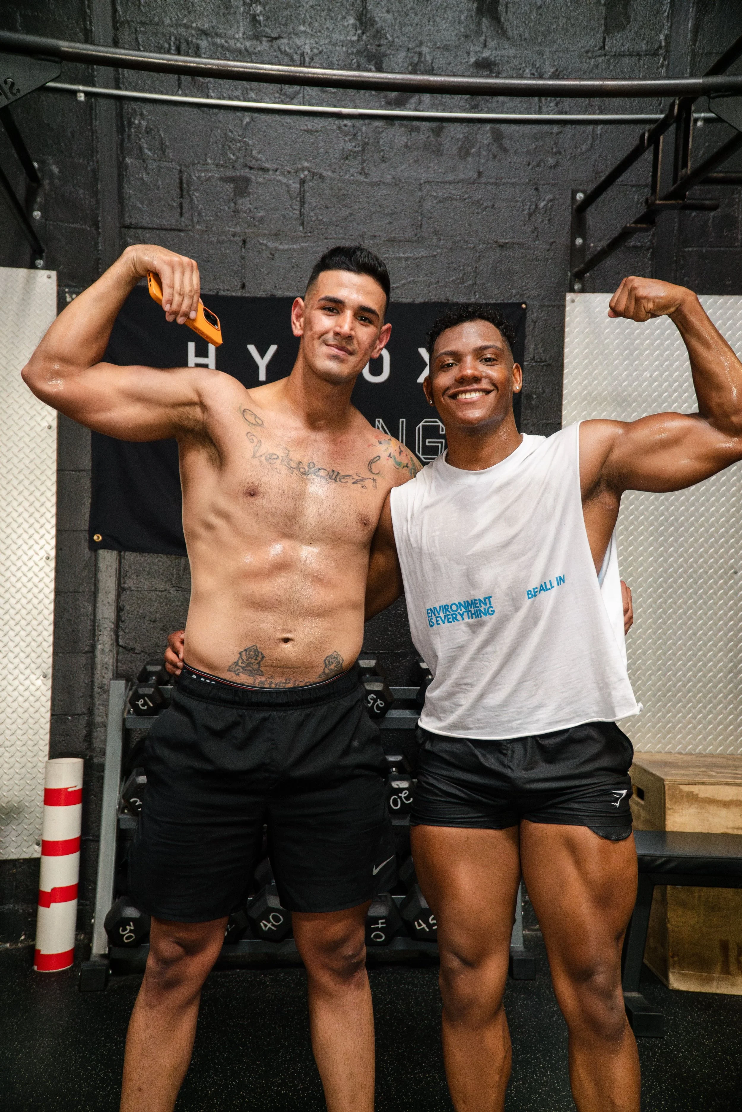 Two muscular men posing in a gym, flexing their biceps. The man on the left is shirtless with tattoos, wearing black shorts, and the man on the right is wearing a white tank top and black shorts, both smiling.