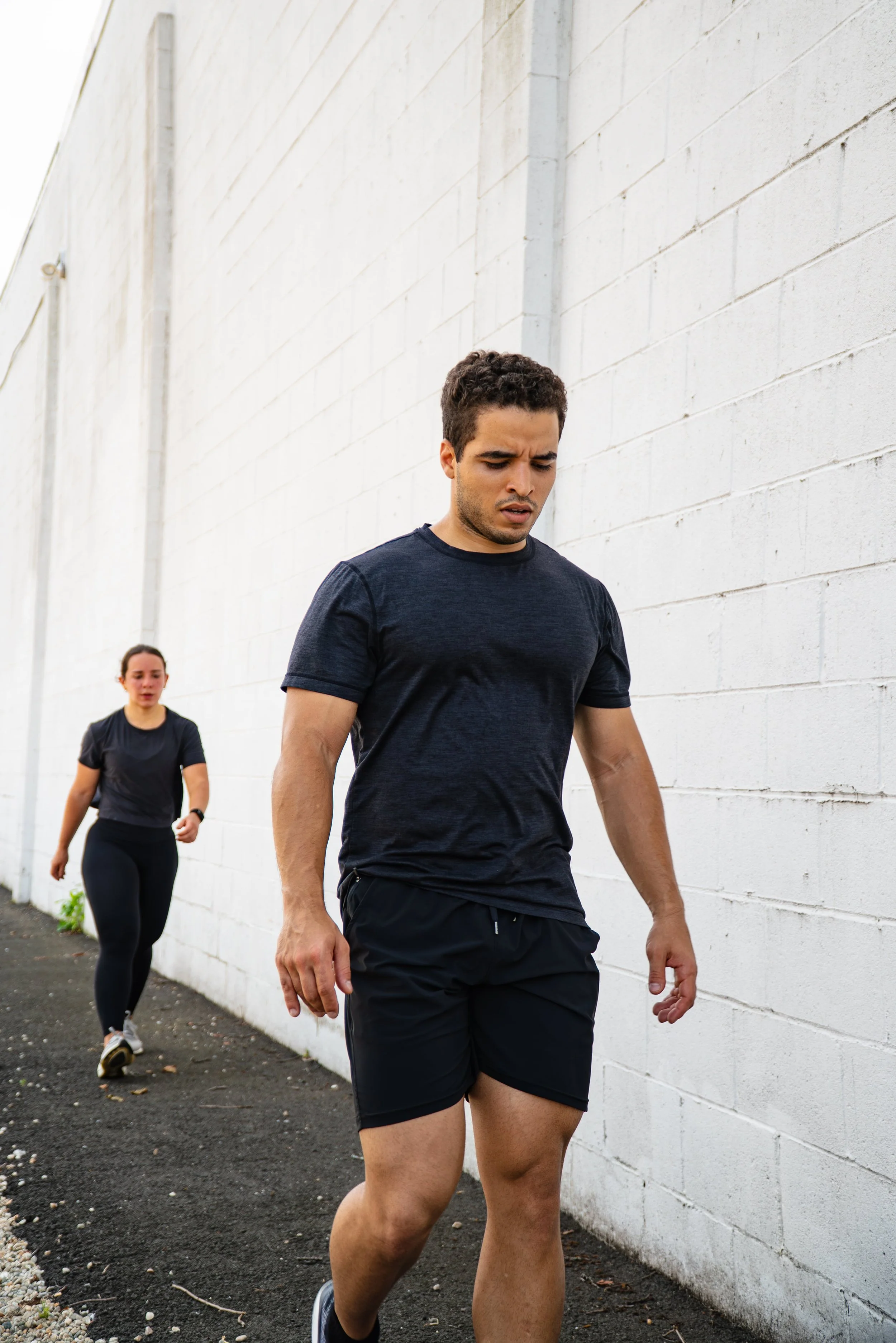 A man and a woman walking outdoors against a white brick wall, dressed in athletic clothing.