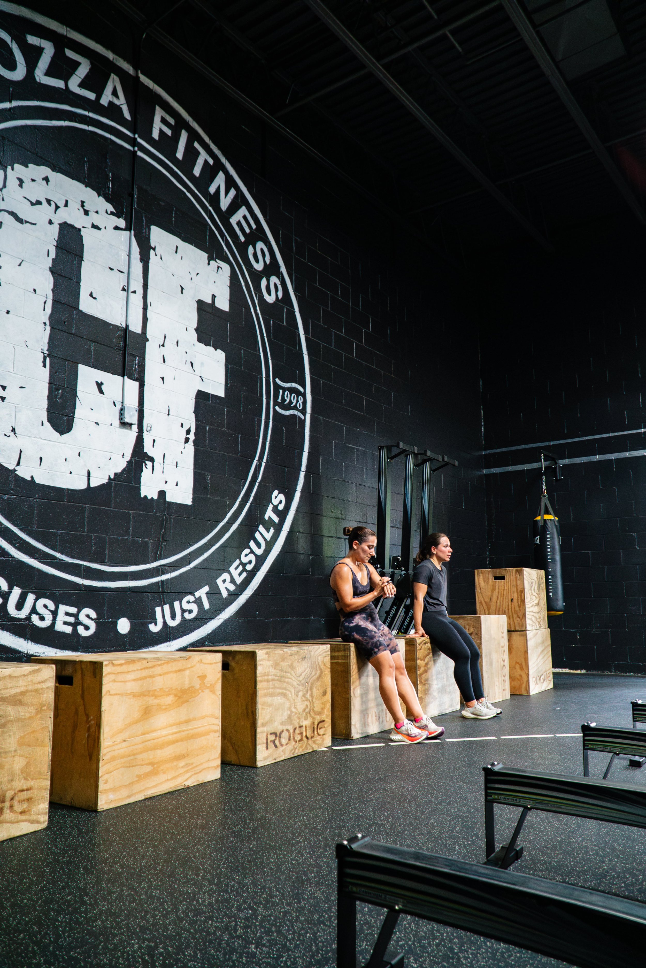 Two women sit on wooden plyometric boxes in a gym, resting after exercise. The gym has a black wall with a large white logo and motivational text, and fitness equipment including a punching bag and martial arts equipment visible.