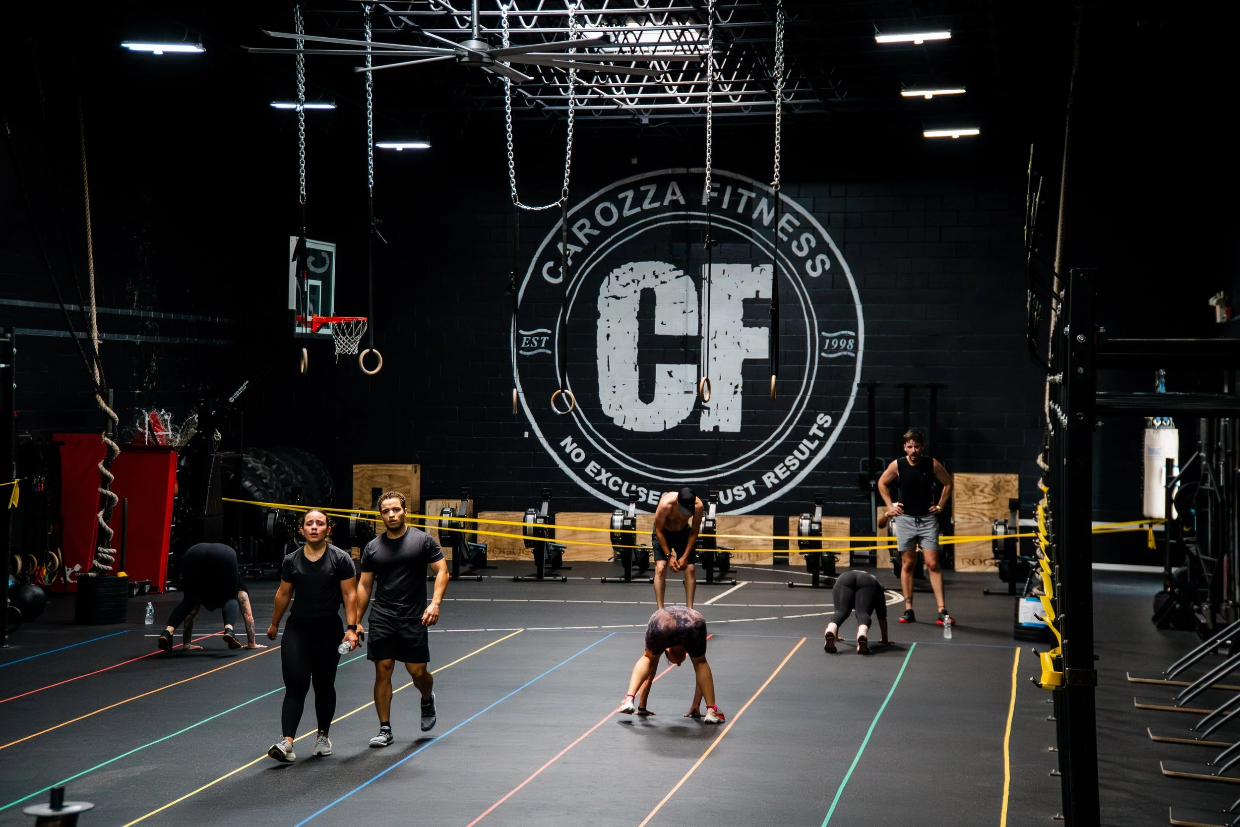 A group of people in a gym space preparing for workout, with some stretching and others adjusting equipment. The gym has a large logo on the back wall reading 'Cairozza Fitness' and features gymnastic rings, a basketball hoop, and various weightlifti