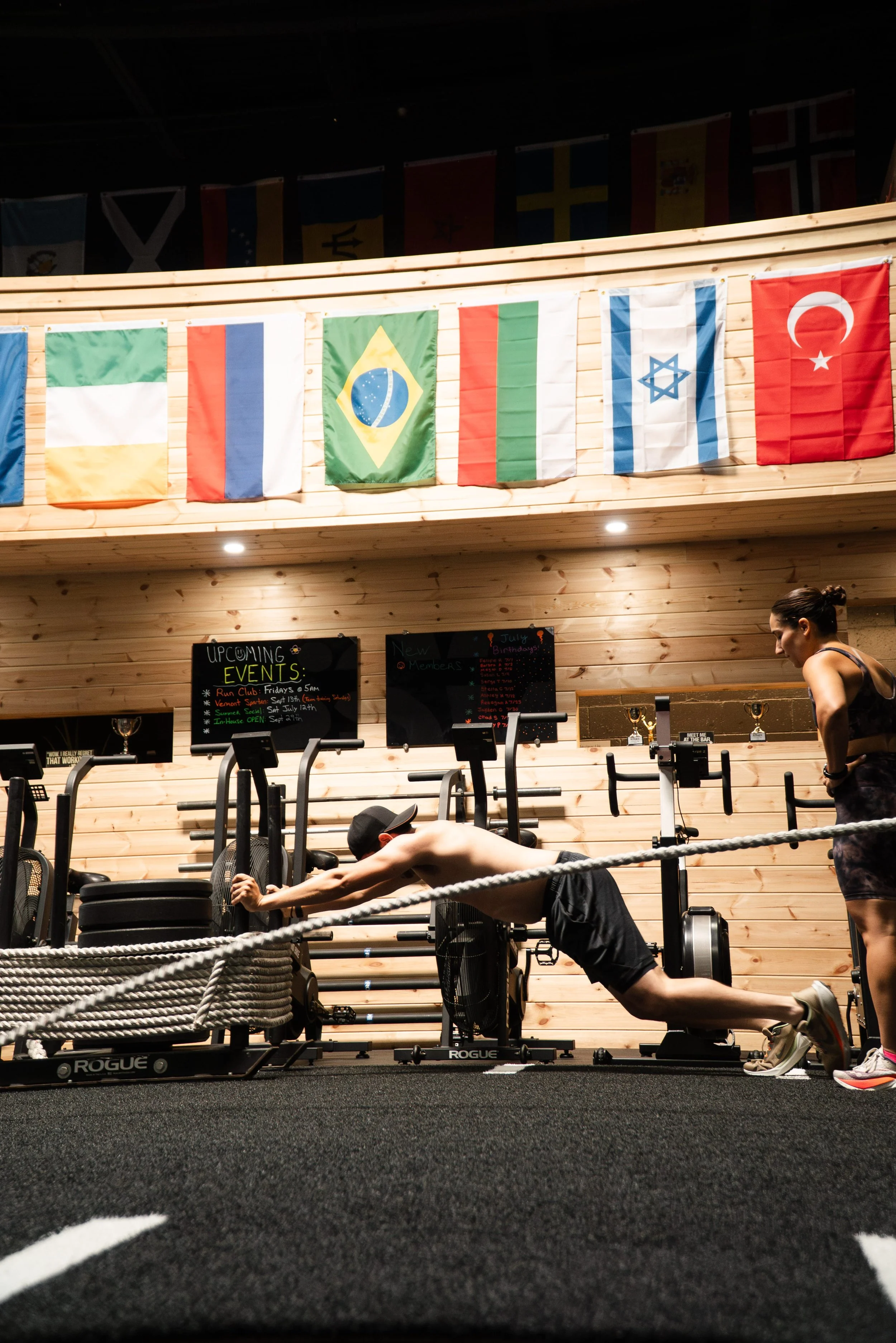 Person exercising on a rope in a gym, with flags of various countries hanging above and sports trophies on a shelf in the background.