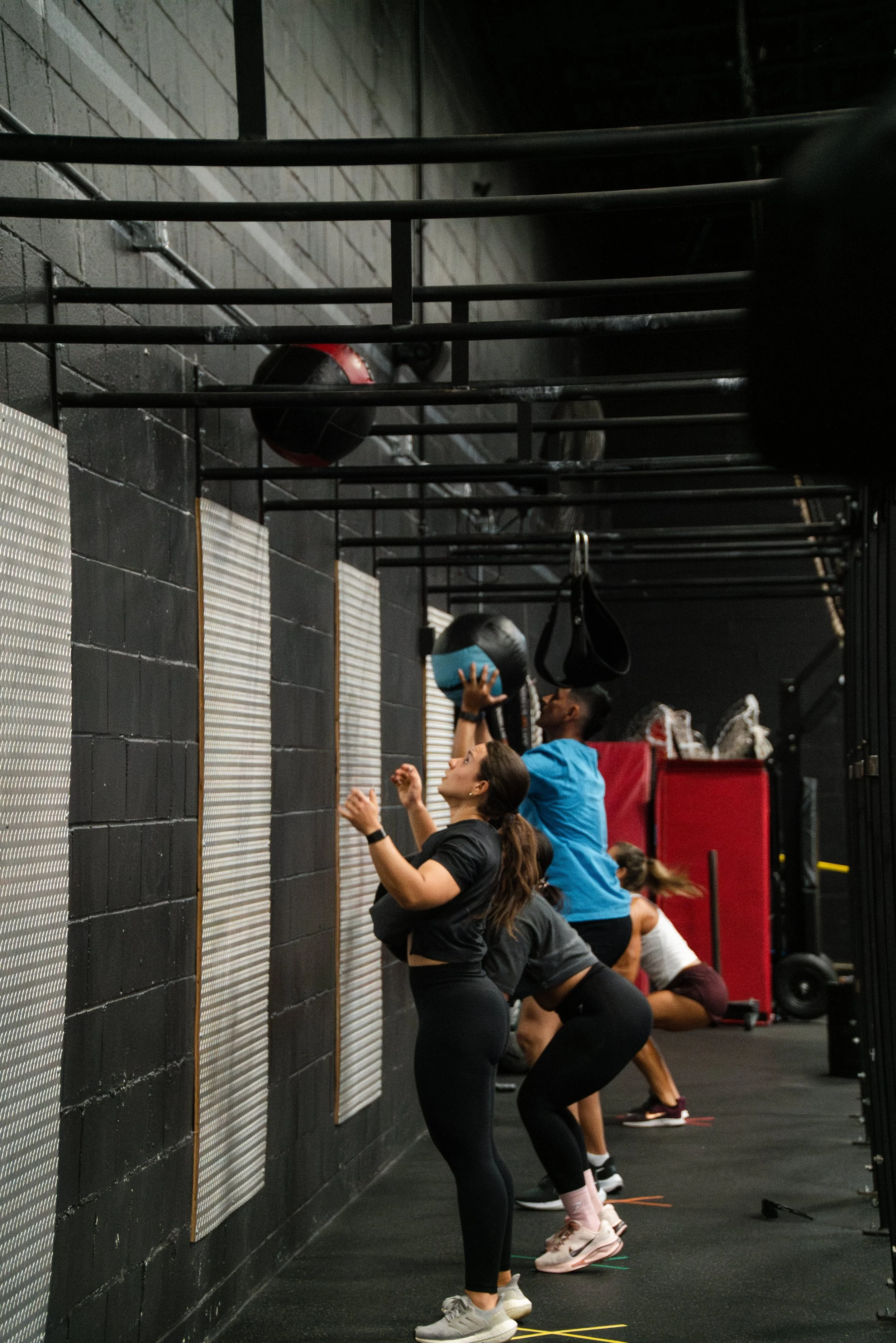 People working out in a gym, performing wall ball exercises with medicine balls against a textured black wall.