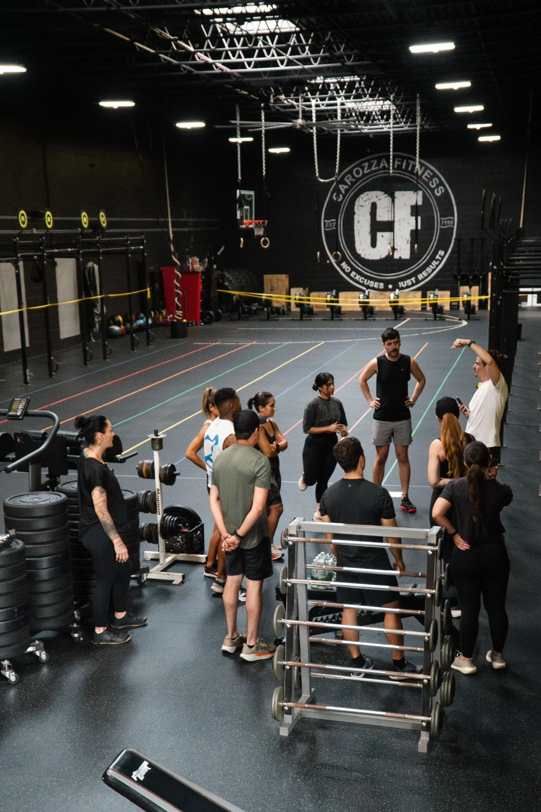 Group of people listening to a trainer in a gym with fitness equipment and a large logo on the wall that reads 'Garozza Fitness CF'.