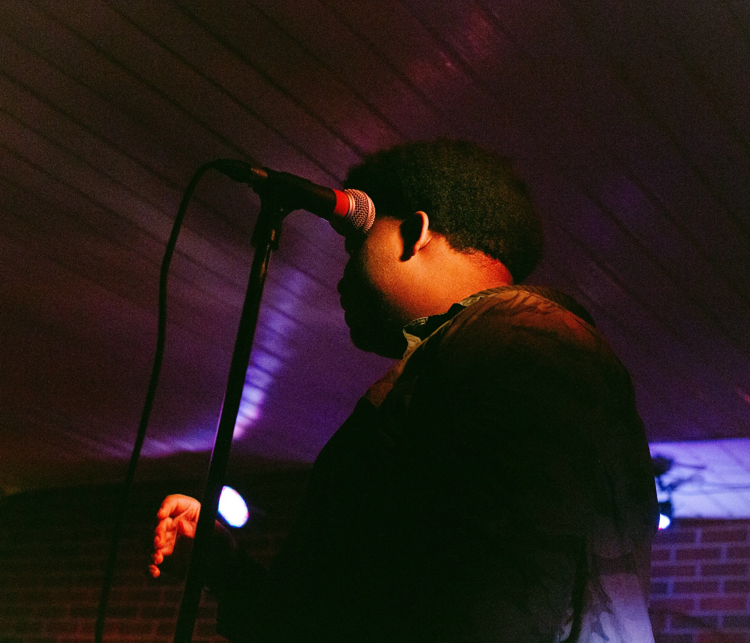 Side profile of man with medium length afro taking a breath away from a microphone. Face and hands highlighted with orange/ gold light. The background is dark, with purple lights. 