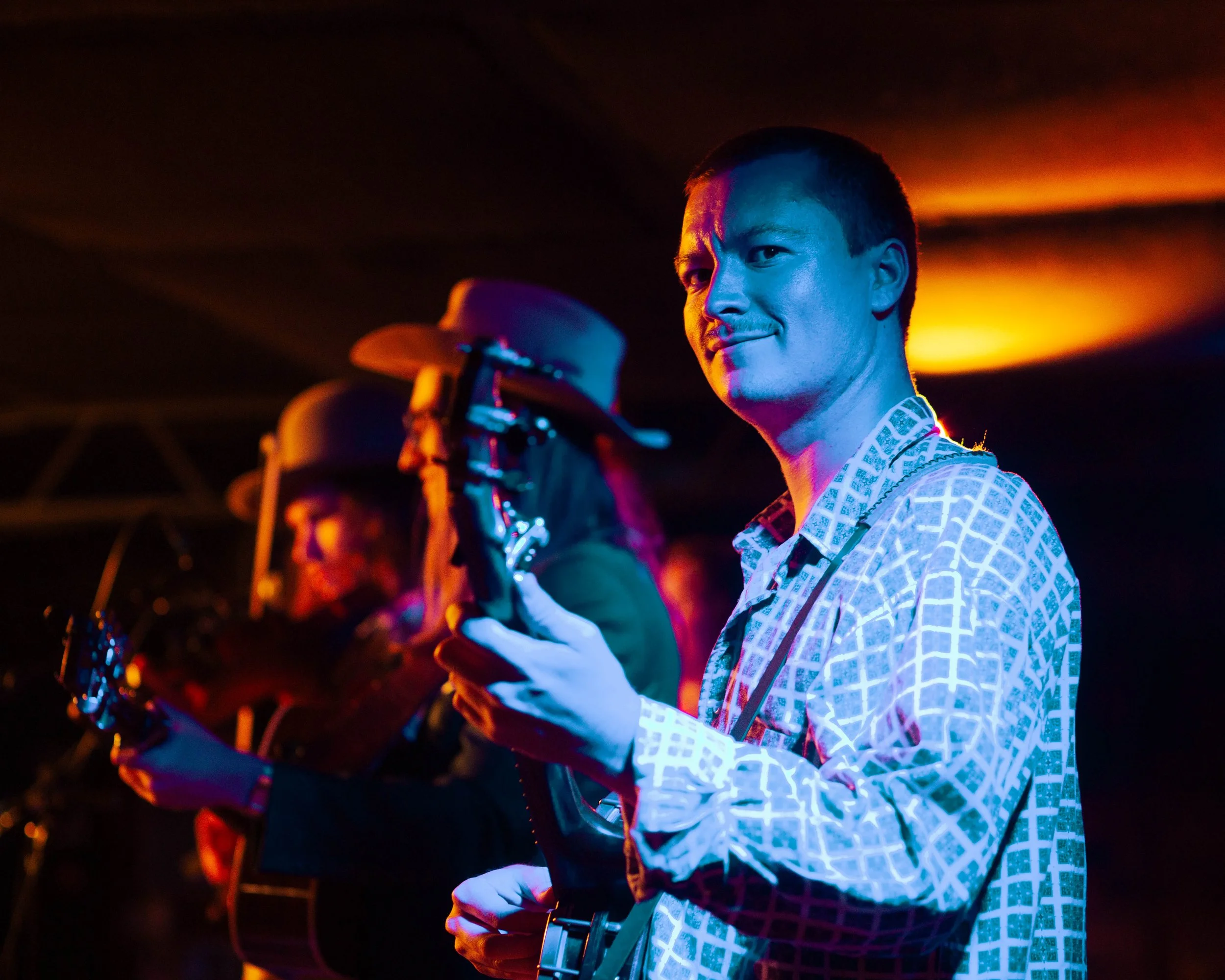 image of 3 bluegrass musicians. Photographed at Station Inn in Nashville. Man in front is playing banjo and cast in blue and orange light.