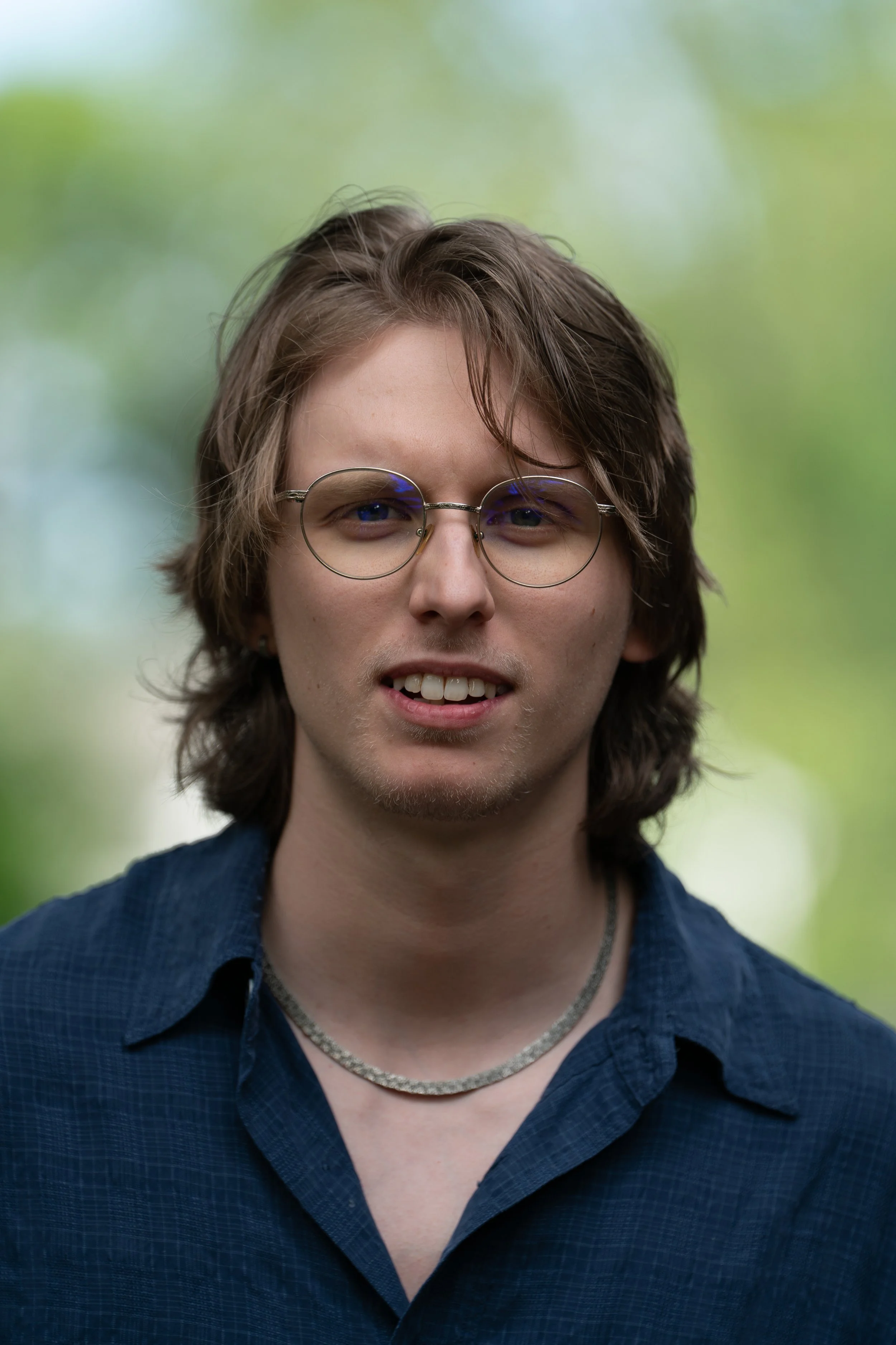headshot portrait of Reed Herring with medium-long brown/blonde hair, wire frame glasses, silver neck chain, blue collared shirt.
