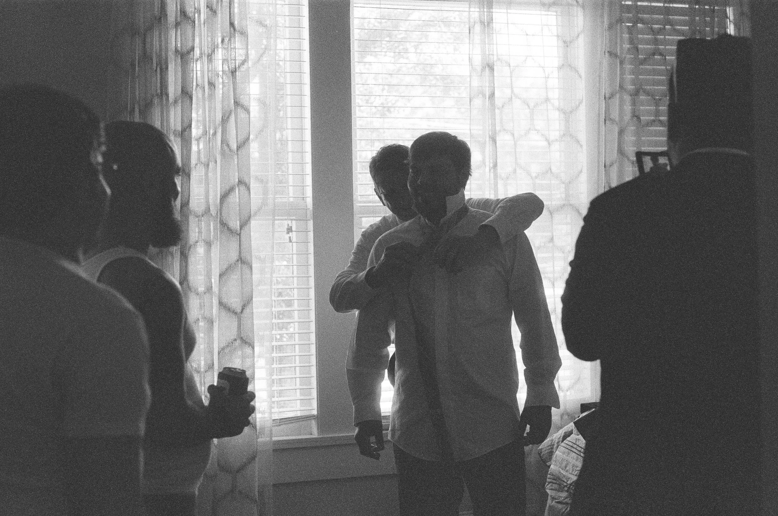 black and white image of father helping son tie necktie from behind. they stand silhouetted before shuttered window. 