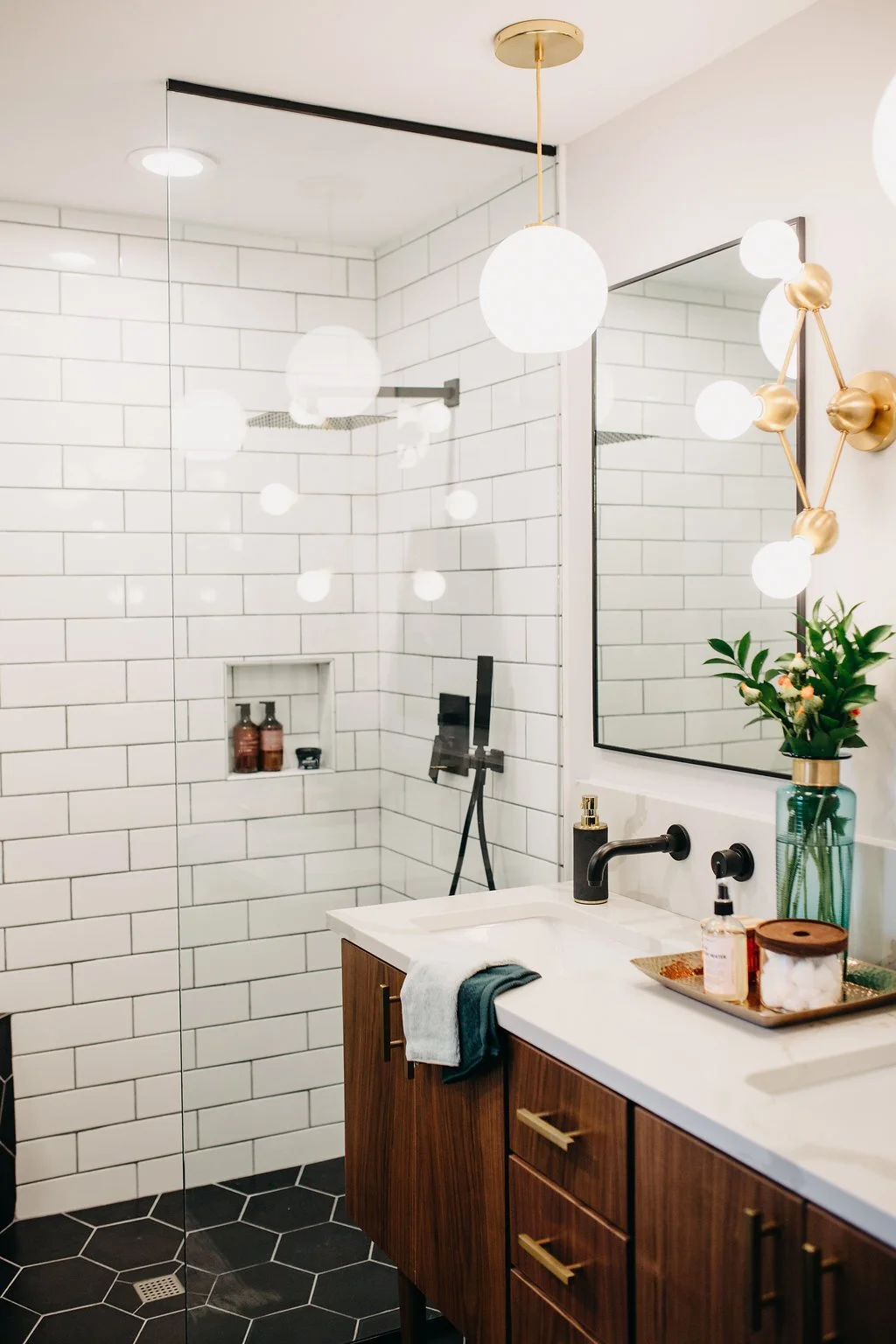 Modern bathroom with white subway tile walls, a glass shower with built-in niche, wooden vanity with a white countertop, a large mirror, and contemporary light fixtures. There is also a green plant on the countertop.