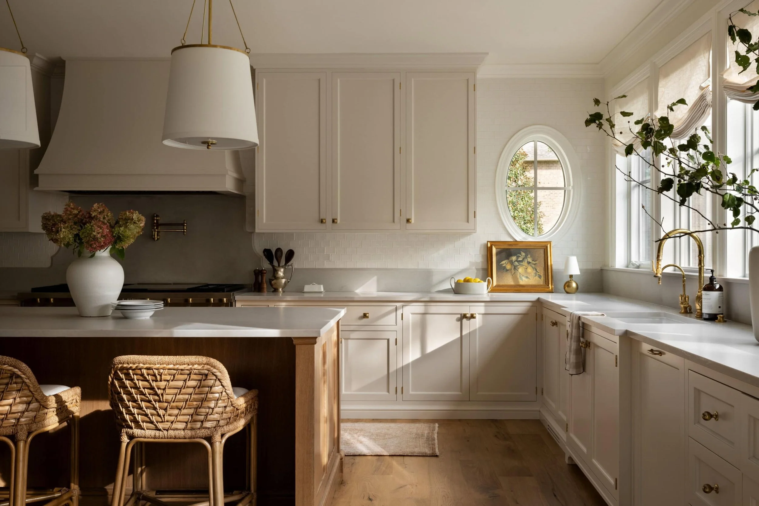 Bright, airy kitchen with white cabinets, a central island with a wood base, woven rattan bar stools, and large windows with beige curtains, decorated with flowers, lemons, and a framed painting.