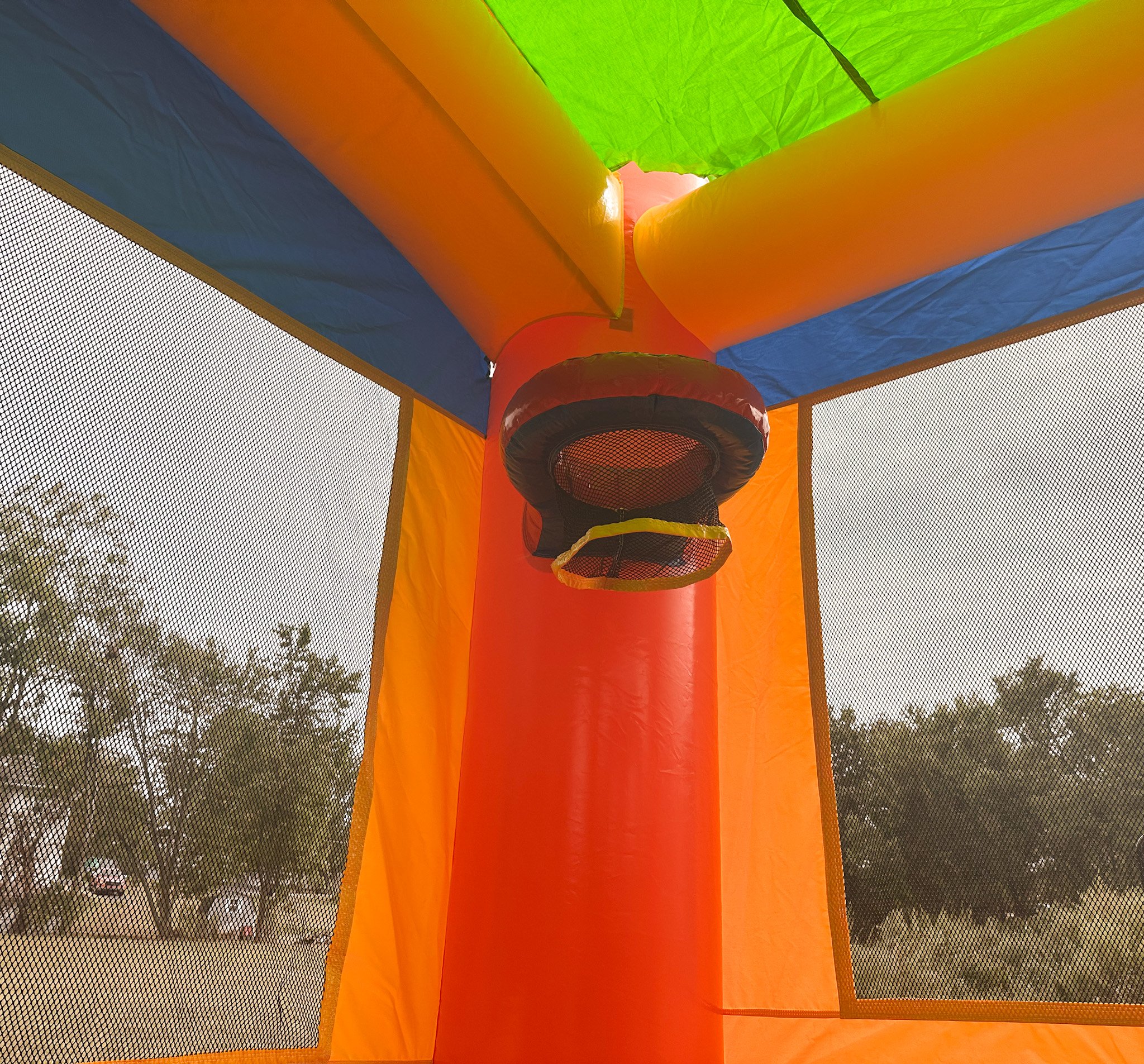 View of the inside of a colorful inflatable bounce house, showing the underside of the ceiling with bright green, orange, and blue sections, and a red inflatable pillar with a black mesh opening.