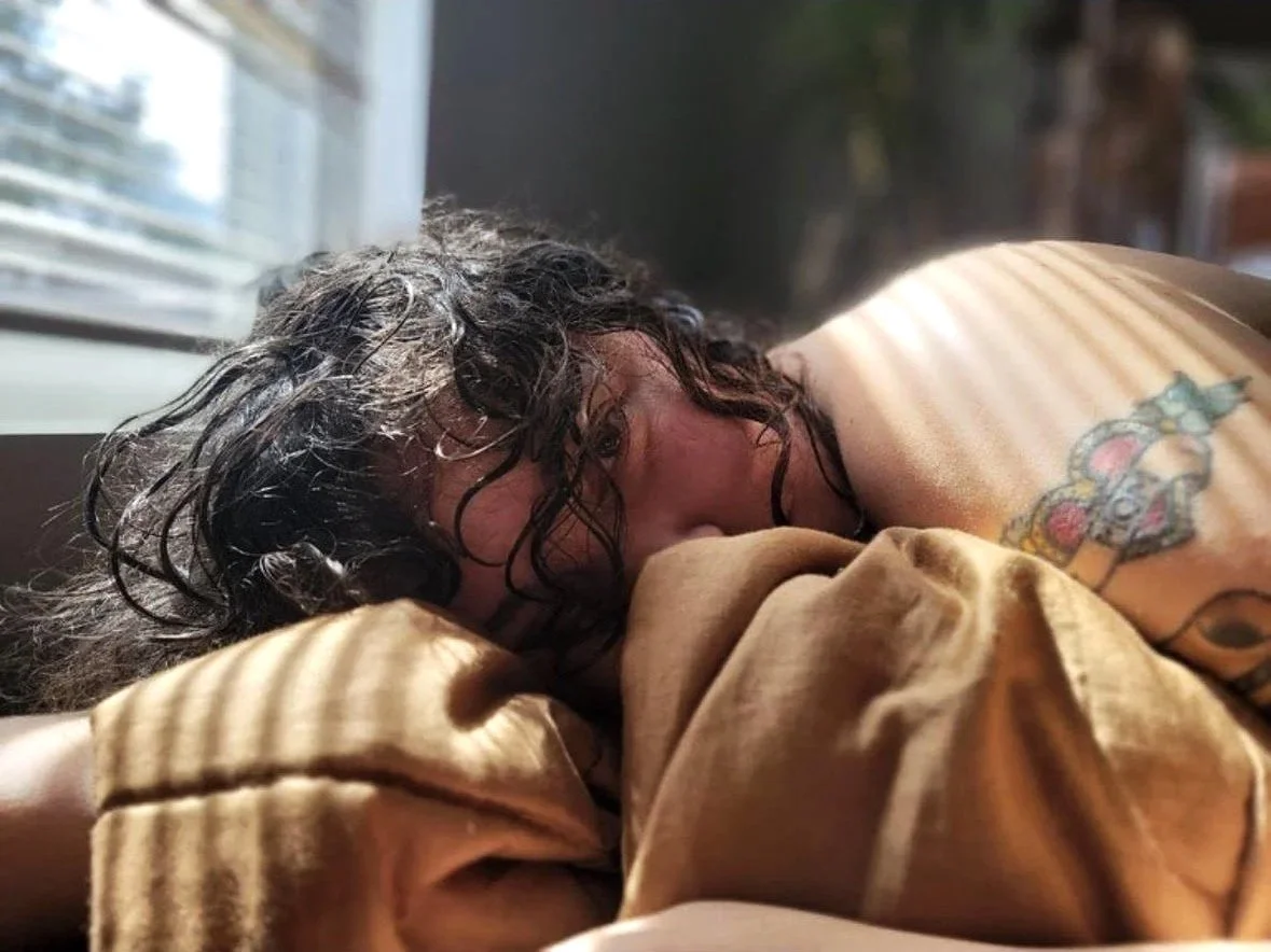 A person with curly dark hair lying on a bed, resting their face on a yellowish-brown blanket, near a window with sunlight casting striped shadows on their shoulder and back, revealing a colorful tattoo on their upper arm.