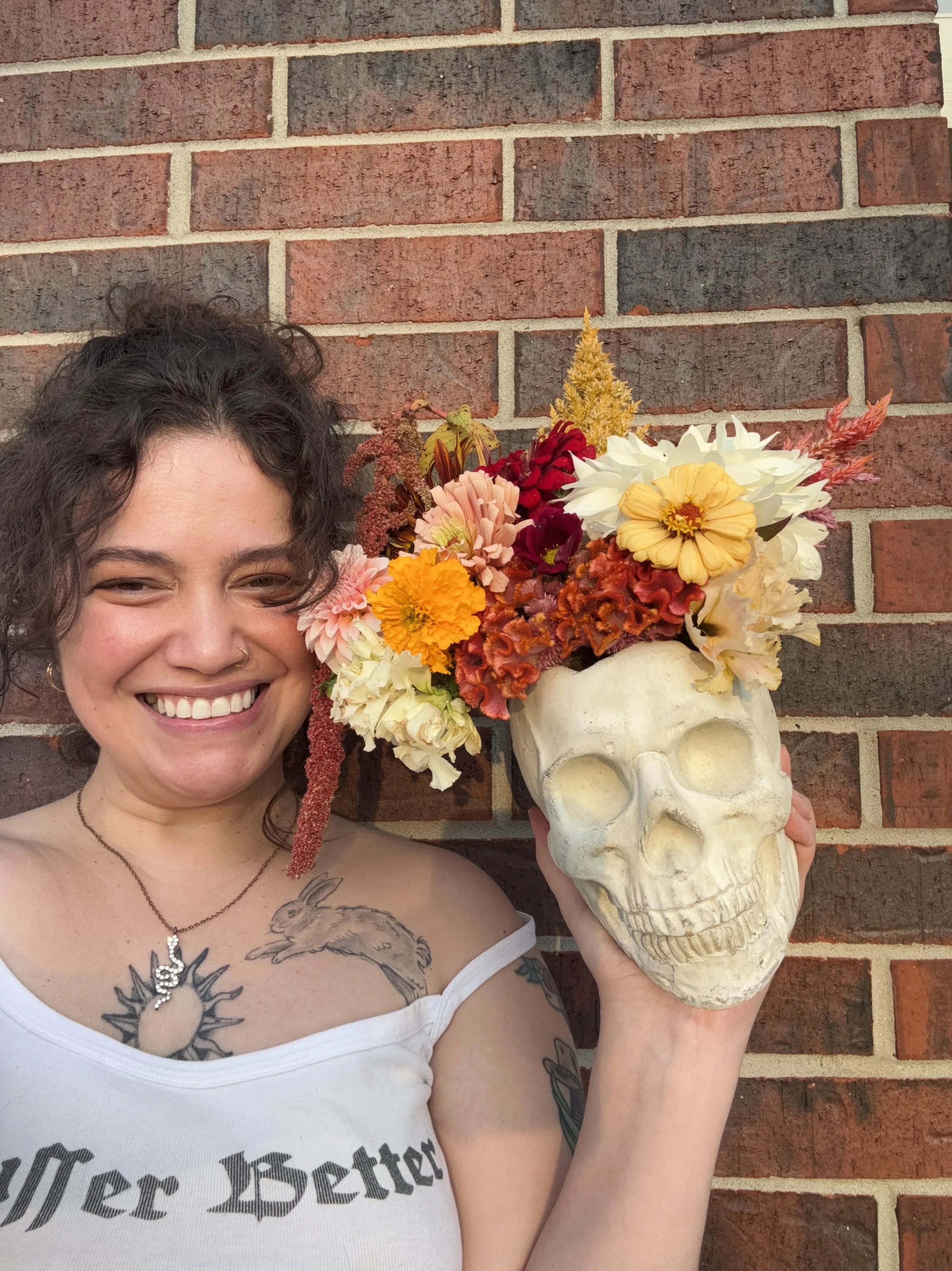 A woman with dark curly hair smiling and holding a skull-shaped vase filled with colorful flowers in front of a brick wall.