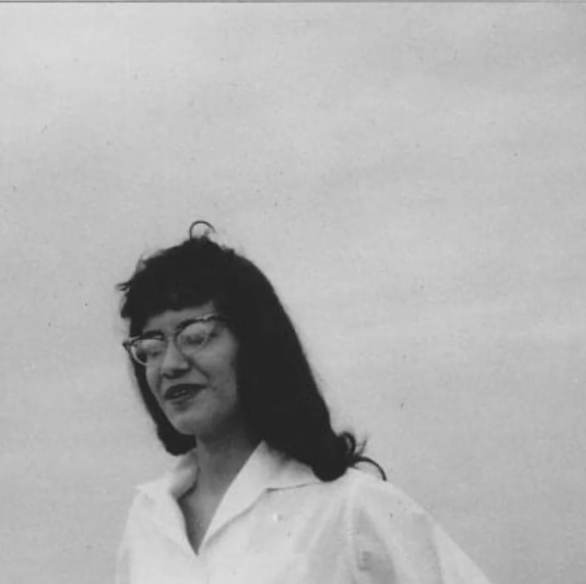 A black-and-white photograph of a woman with dark hair and glasses, wearing a white shirt, against a plain light background.