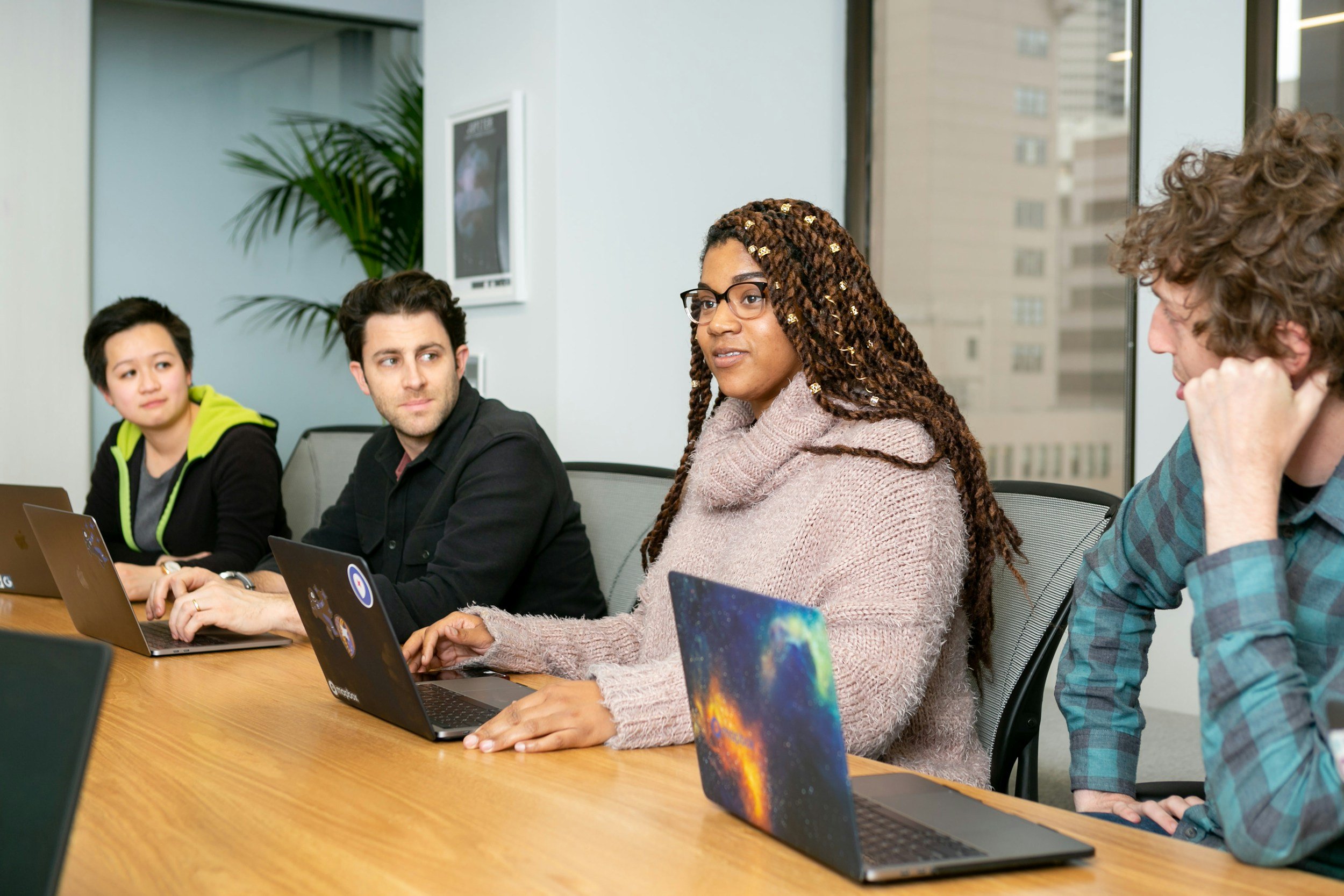 Four diverse individuals seated at a conference table with laptops, engaged in a discussion in a modern office with large windows and a cityscape view.
