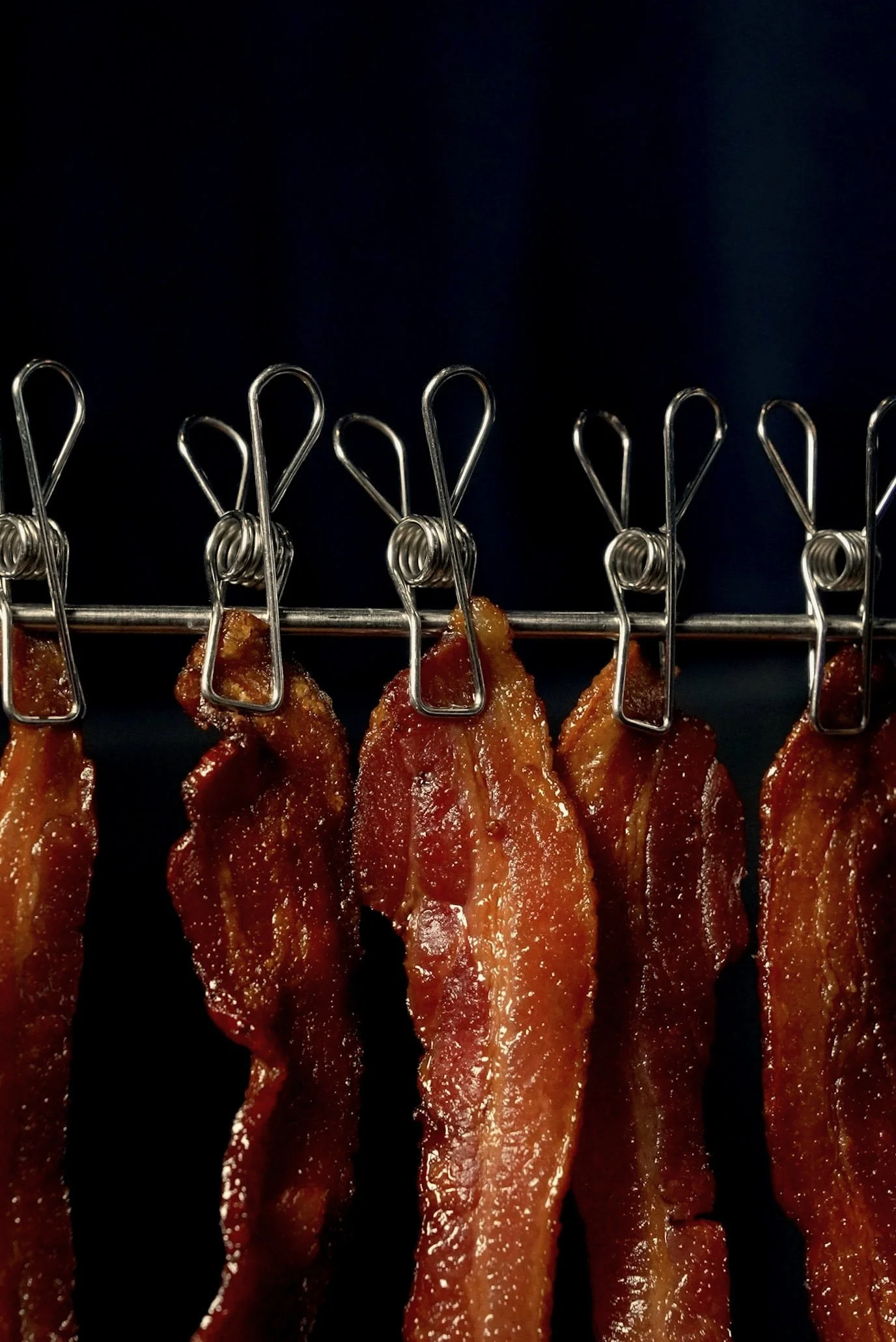 Several strips of cooked bacon hanging from metal hooks on a rack against a dark background.
