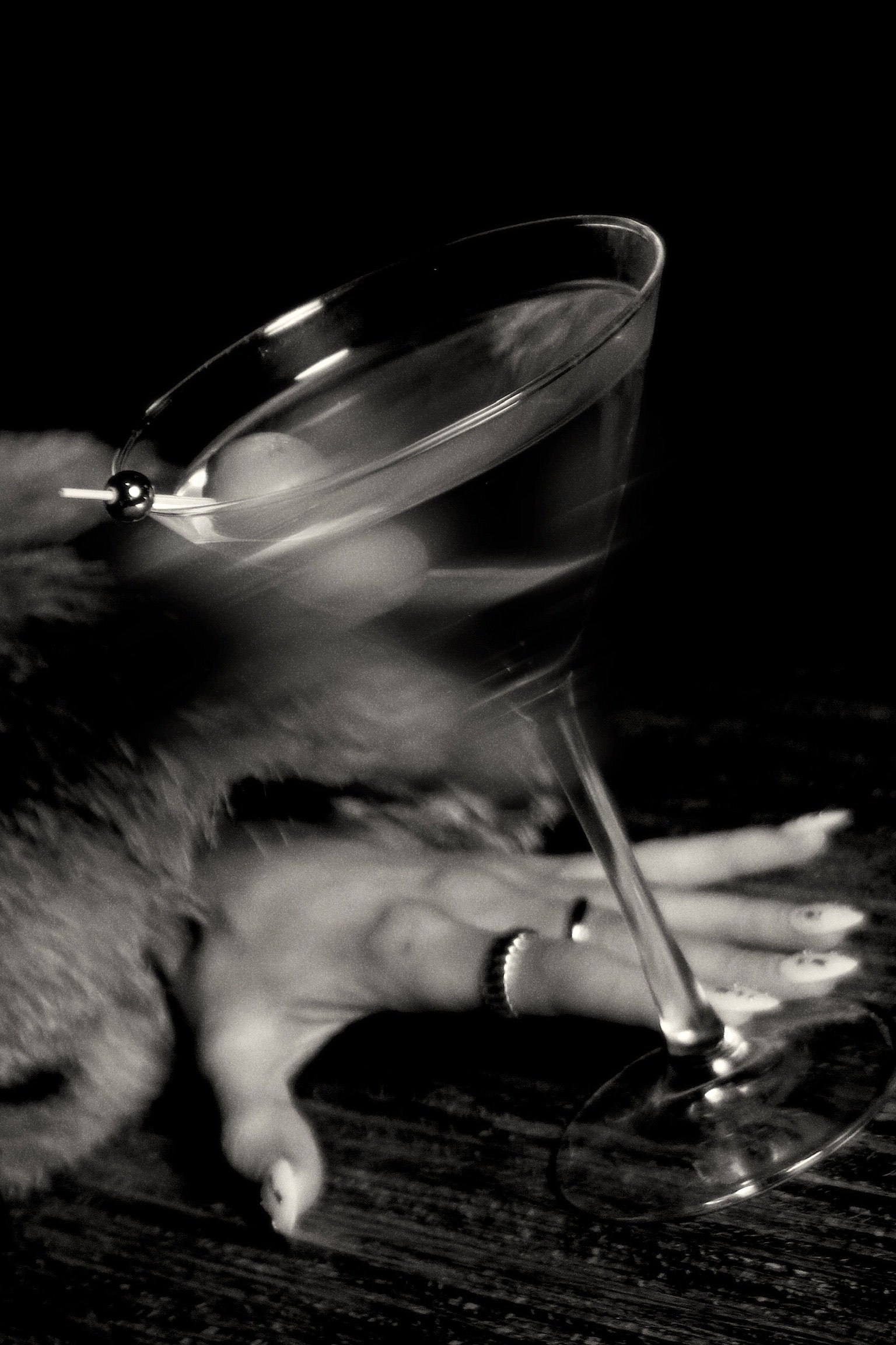 A martini glass with an olive, being filled with a drink, on a table with a smoky or foggy effect, black and white photo.