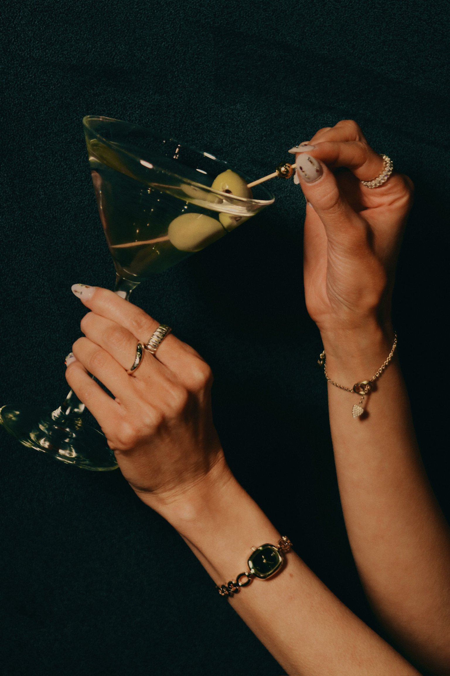 Close-up of hands holding a martini glass with olives. The hands are adorned with rings and bracelets. The background is dark.