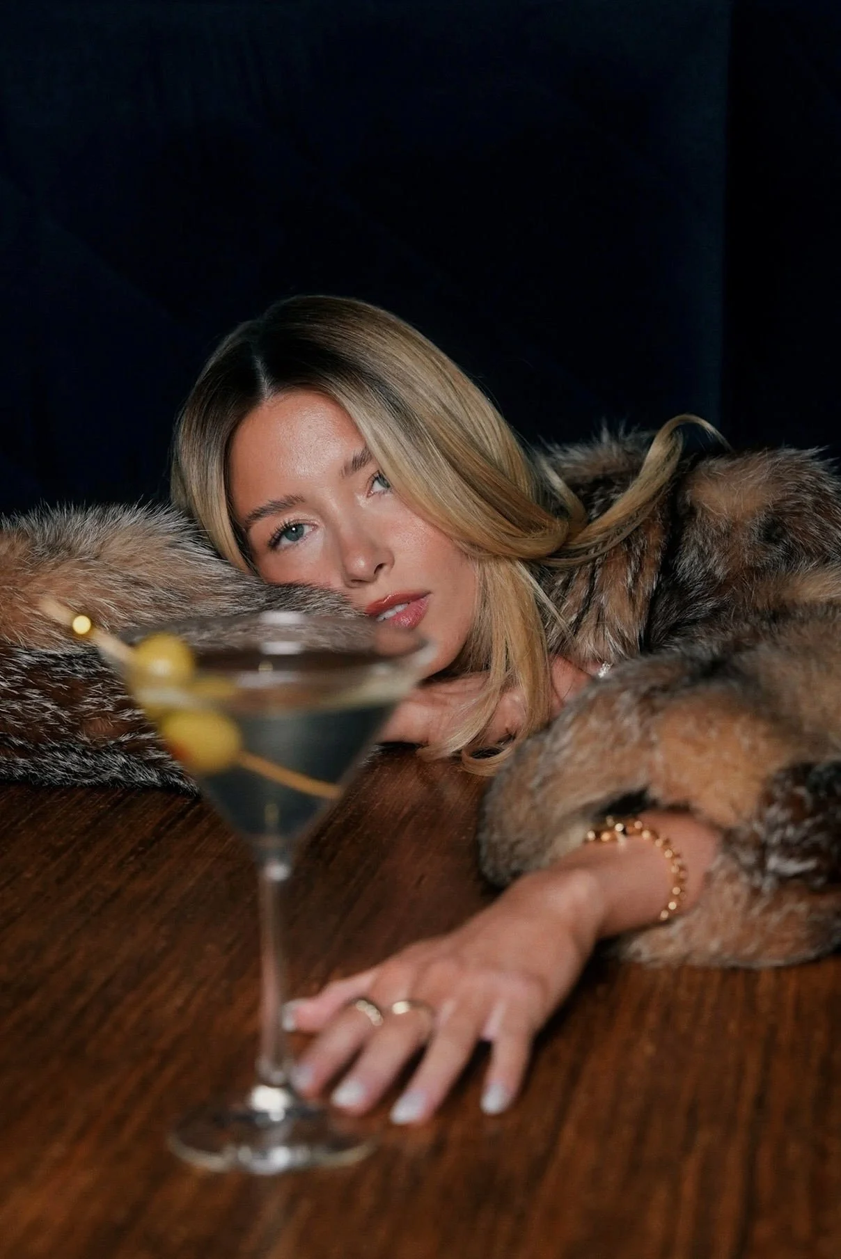 A woman with blonde hair lying on a wooden table, wearing a fur coat, with her head resting on her arms near a cocktail glass with olives, and gazing into the camera.