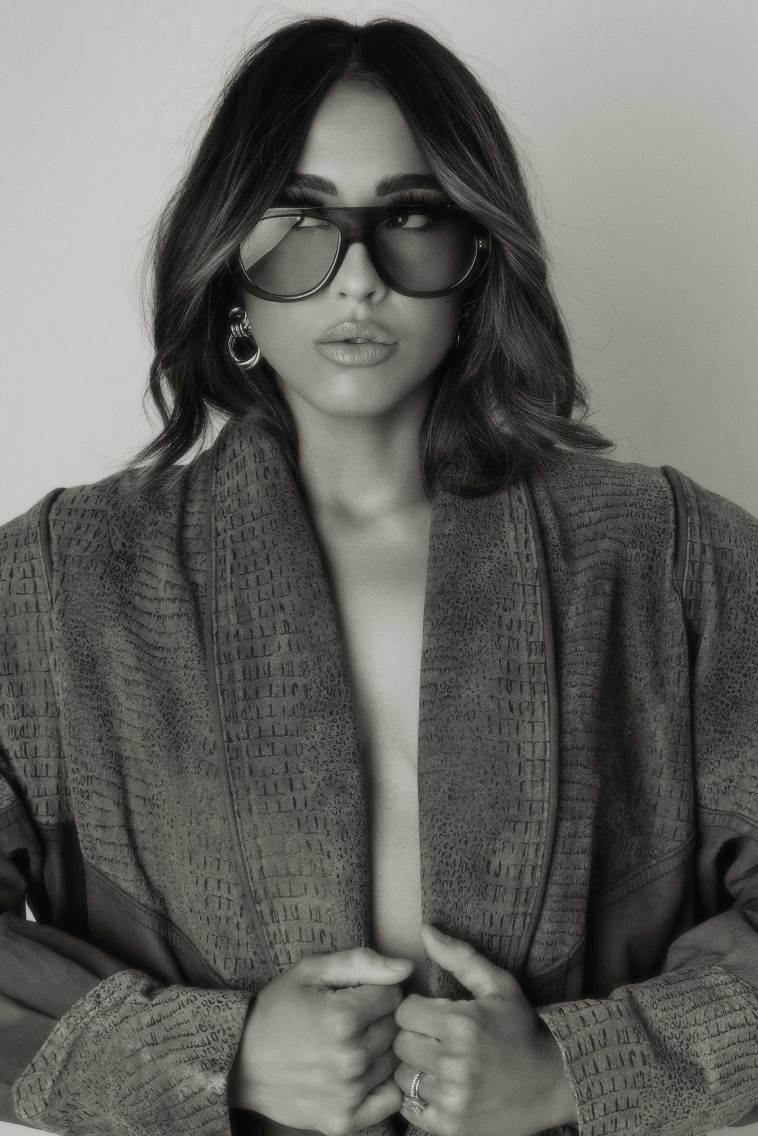 A woman with dark hair wearing large sunglasses and earrings, dressed in a textured jacket, posing with her hands clasped together against a plain background.