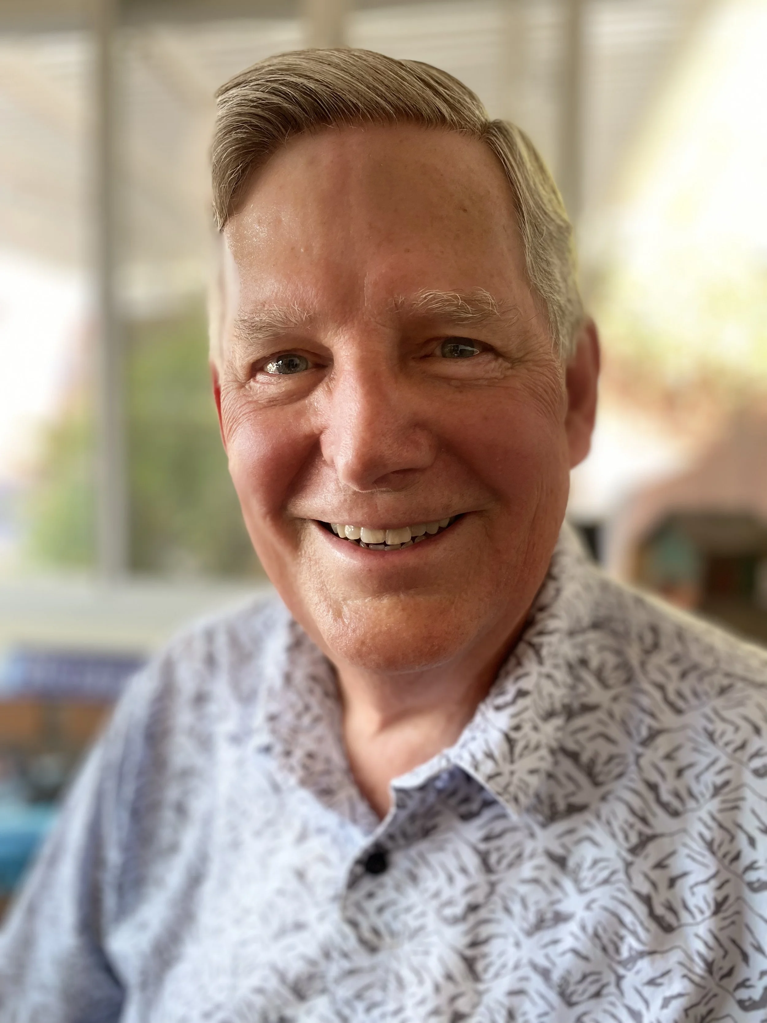 Close-up of smiling middle-aged man with short blond hair, wearing a white patterned shirt, in front of blurred background with windows and natural light.