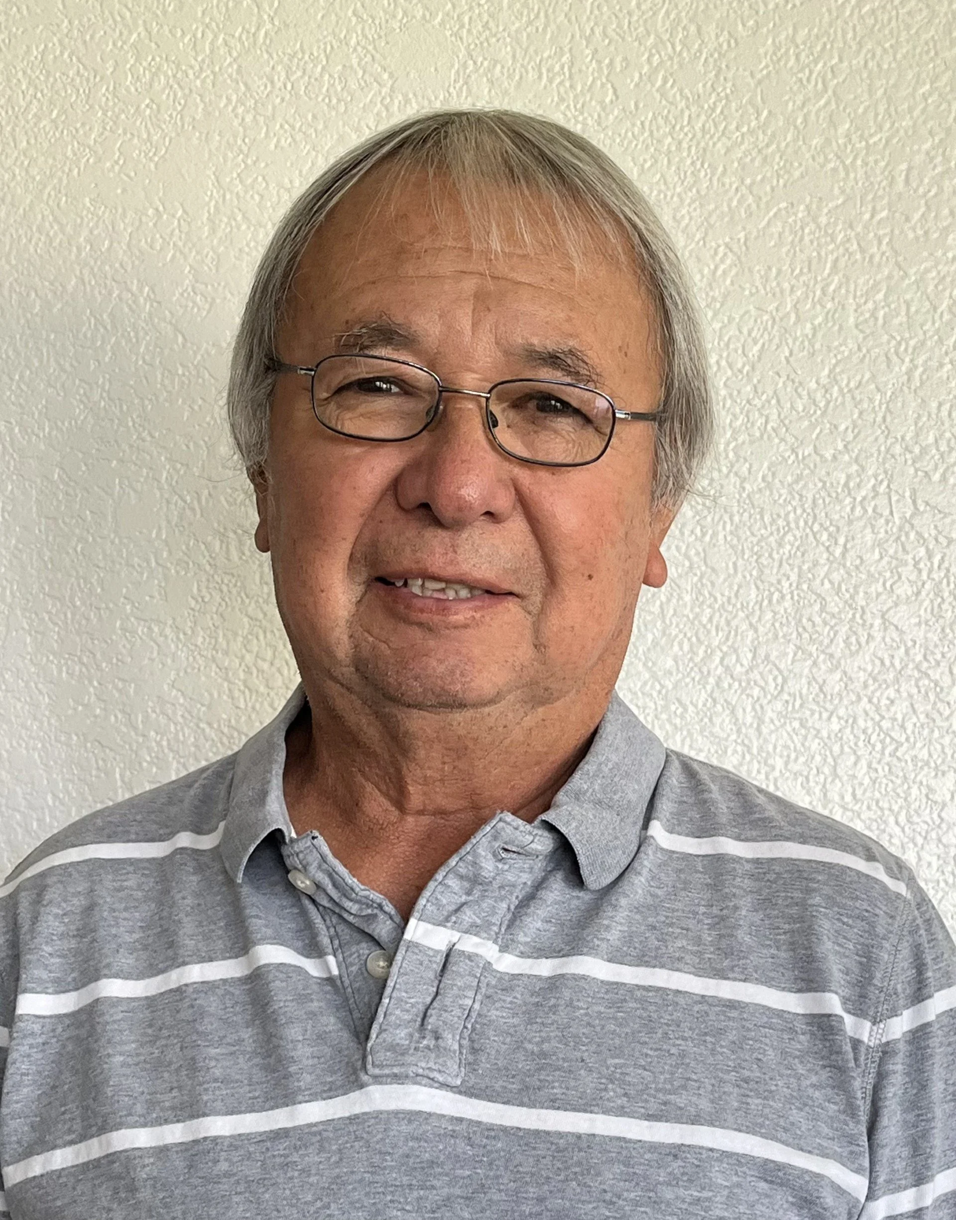 A middle-aged man with gray hair and glasses smiling at the camera, wearing a gray and white striped polo shirt, standing against a textured beige wall.