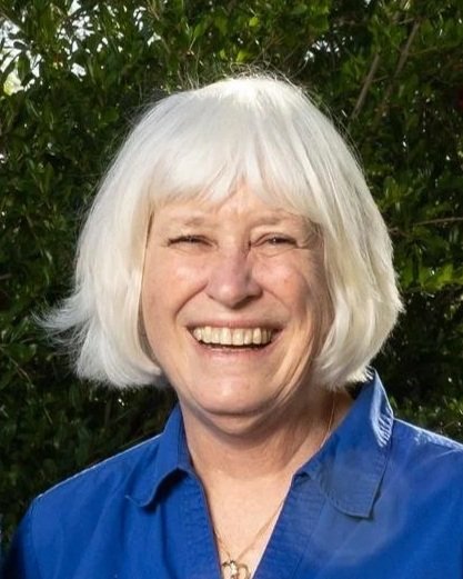 A woman with white hair smiling outdoors with green foliage in the background.