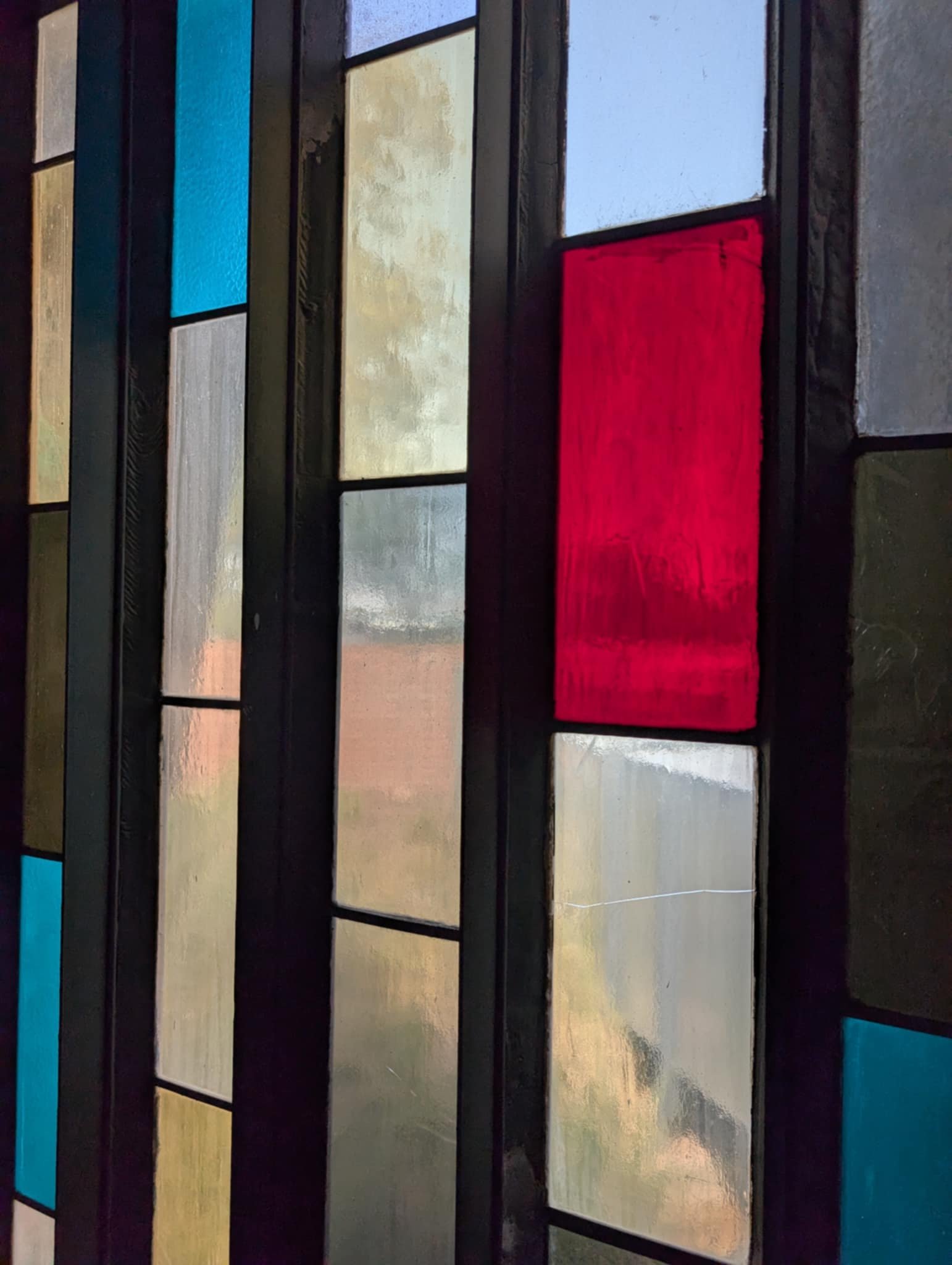 Close-up of a colorful stained glass window with rectangular and square glass panes in red, blue, yellow, clear, and black, with a metal frame.
