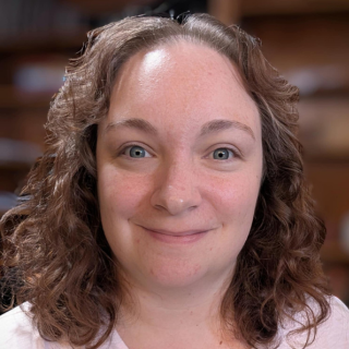 A woman with curly brown hair and blue eyes smiling at the camera in an indoor setting.