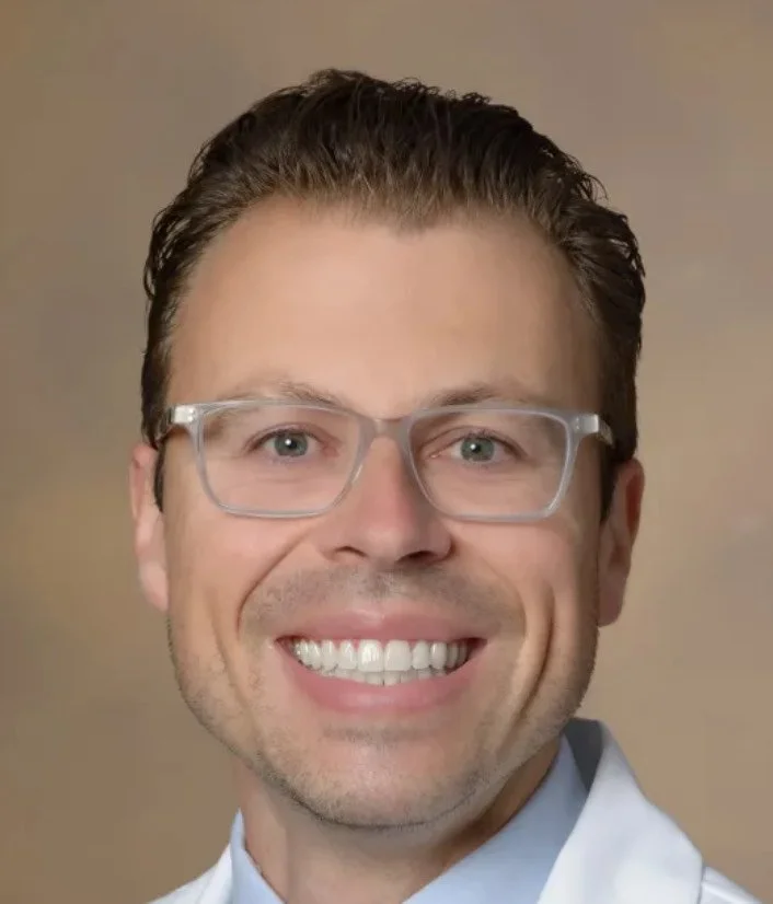 Close-up portrait of a smiling man with glasses and brown hair, wearing a white shirt.