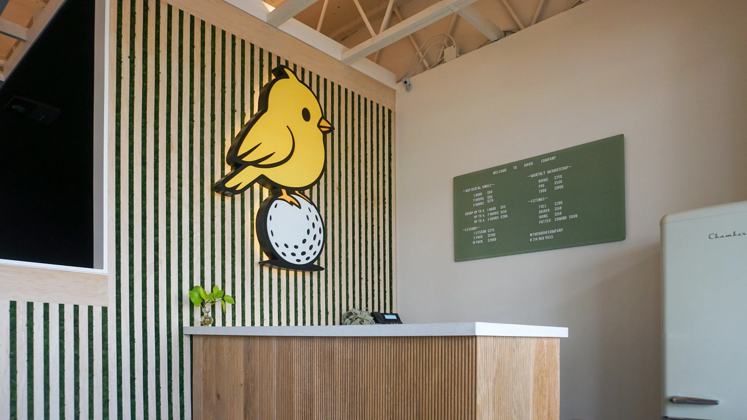 Interior of a modern space with a yellow bird sign on the wall, a green menu board, a small plant on a white countertop, and a white refrigerator.