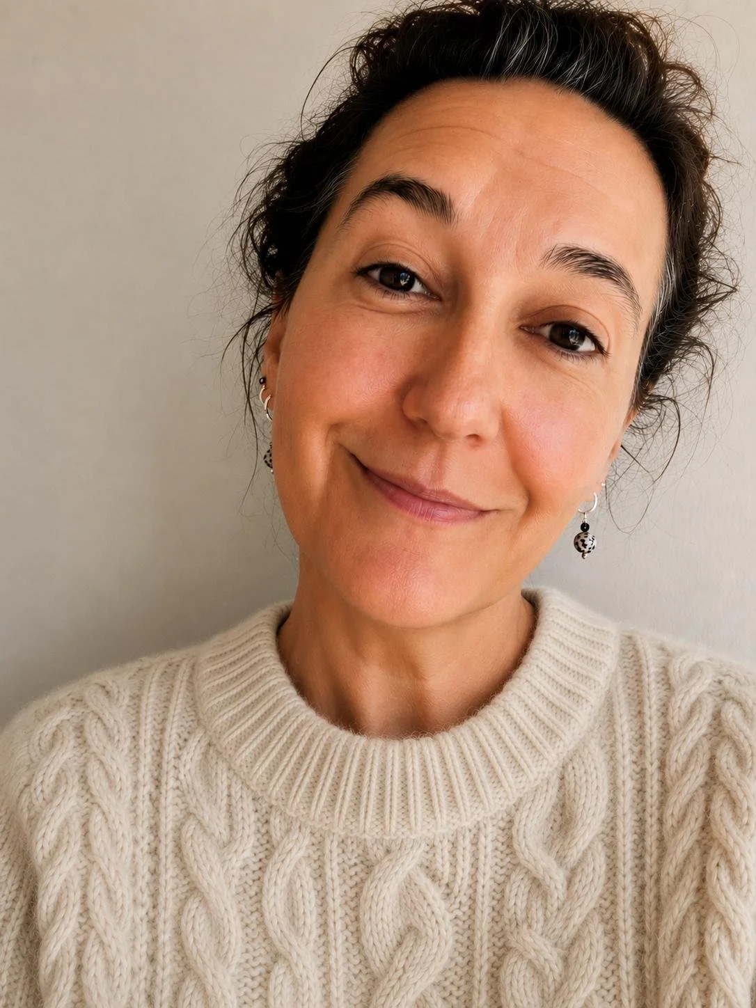 Maternity nurse and life coach for women Solange Martins is wearing a cream-colored cable-knit sweater against a plain wall background.