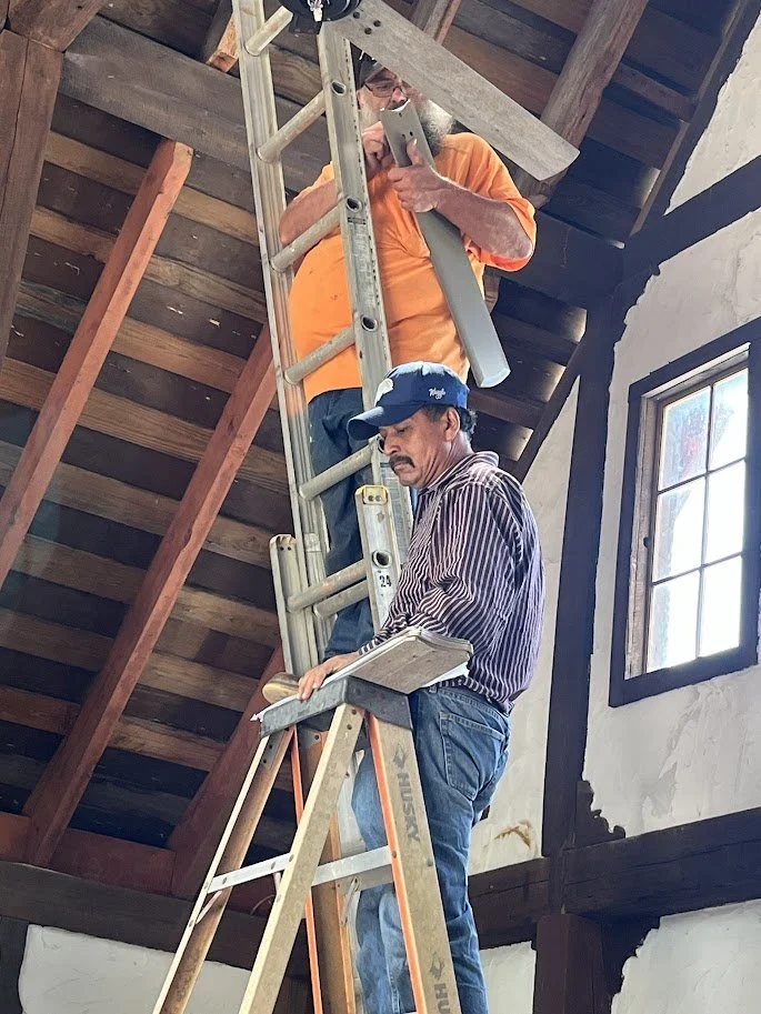 Two men working on a ladder inside a room with an unfinished wall and exposed wooden beams ceiling.