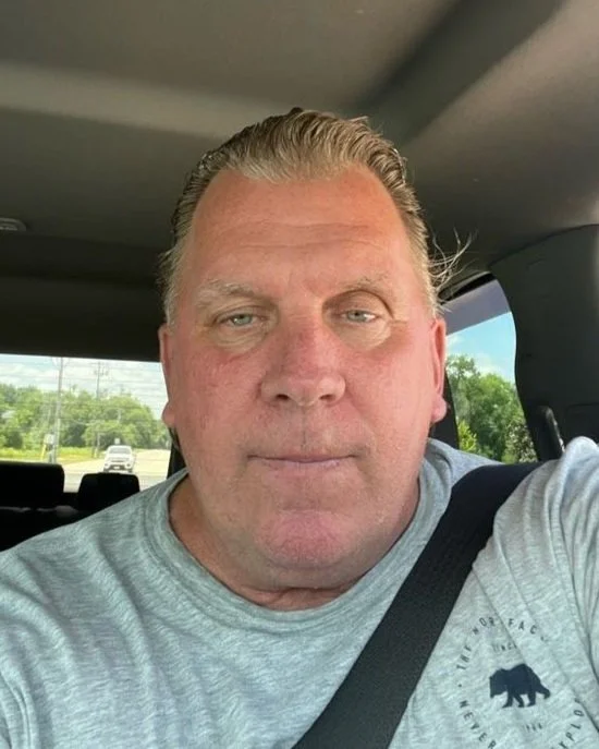 A middle-aged man with light skin and slicked-back blond hair taking a selfie inside a vehicle, with a seatbelt across his chest. The background shows a highway with trees and cars.