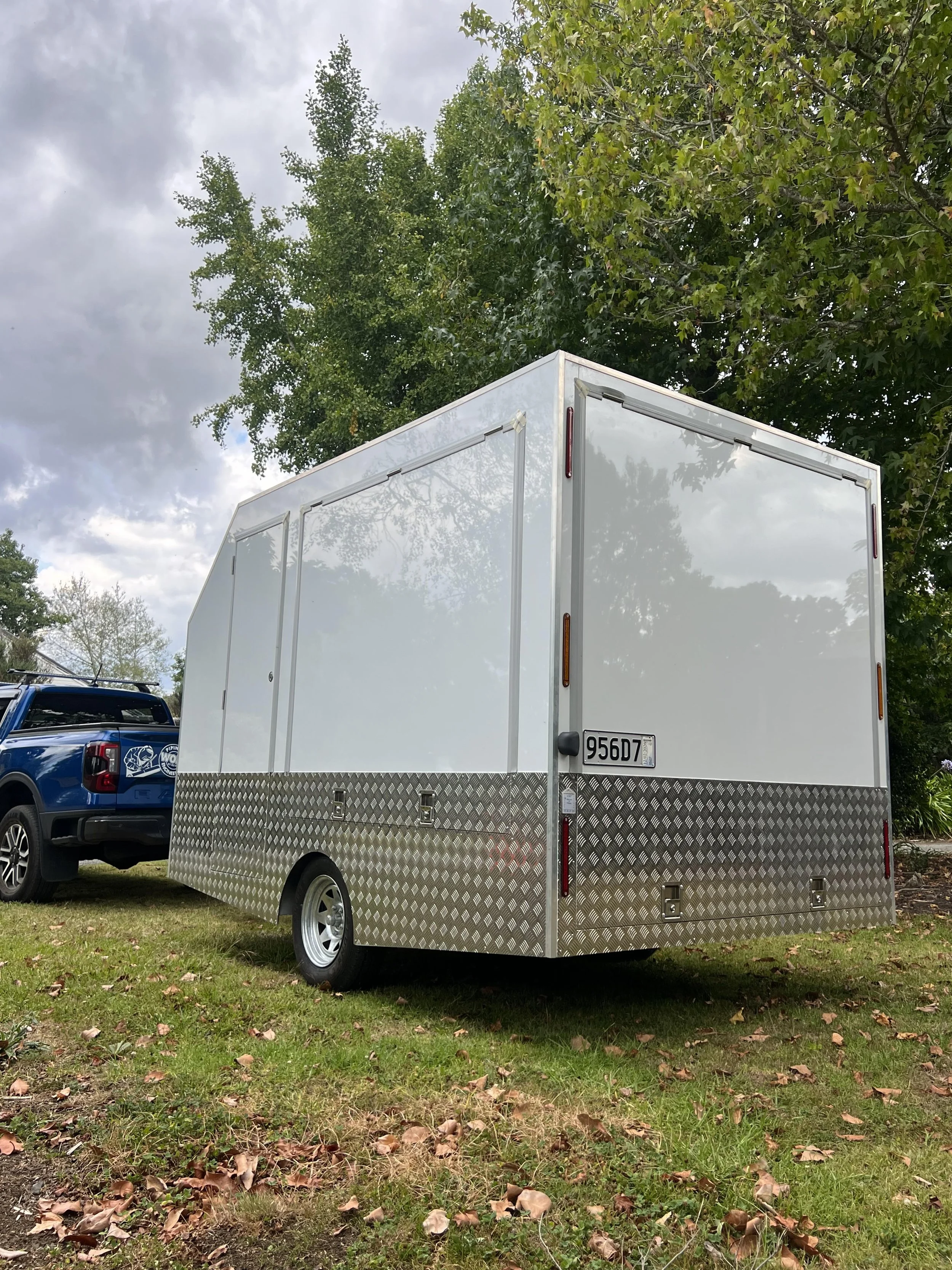A white utility trailer with diamond plate metal trim parked on grass, with a blue pickup truck and trees in the background.