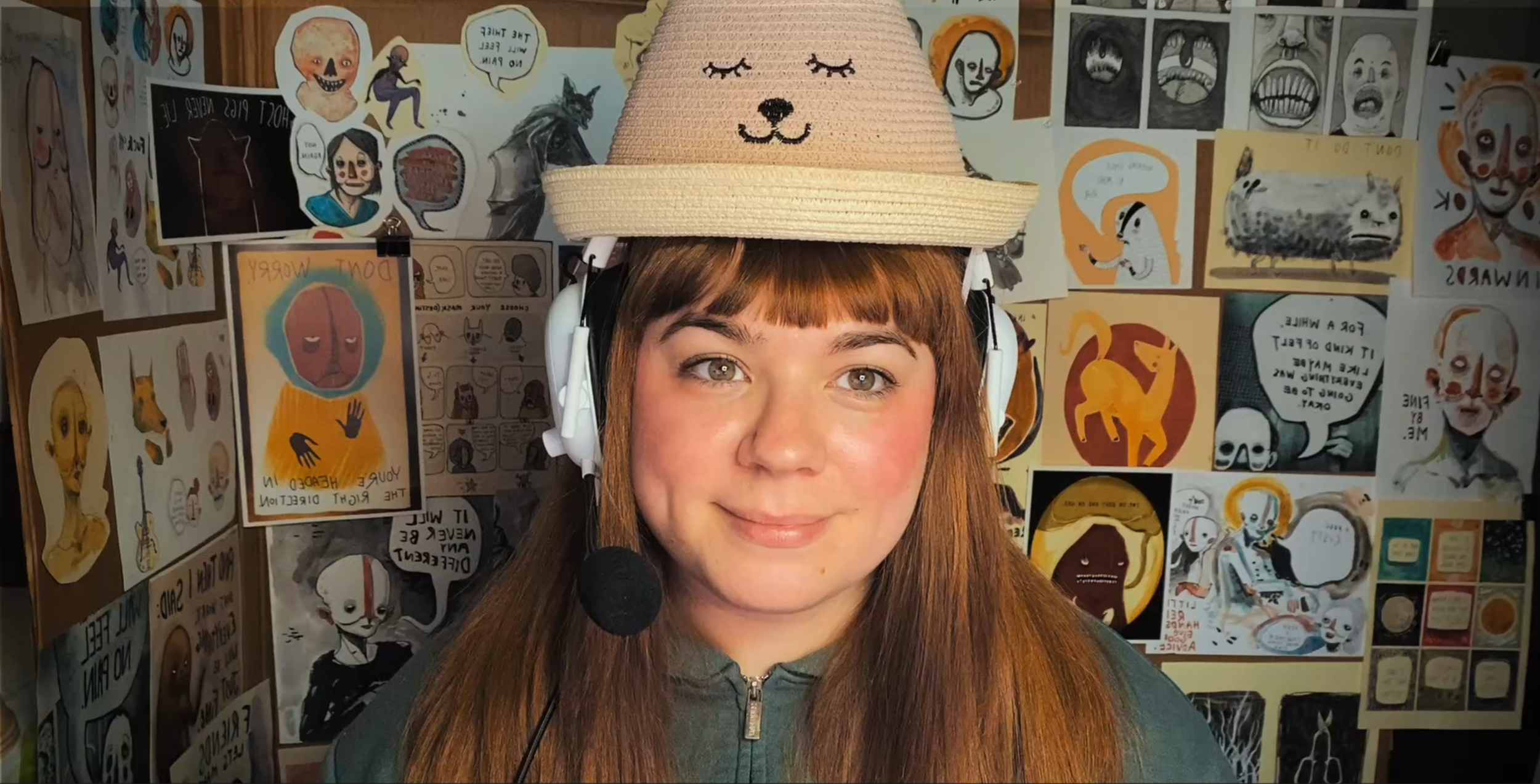 A woman with long hair wearing a hat with a cute cat face on it, along with headphones and a microphone attached to her cheek, is smiling at the camera. The background is filled with various illustrated posters and drawings, many featuring cartoon-like characters with faces.