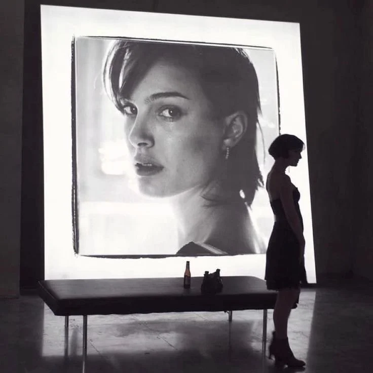 A woman in profile walking past a large black-and-white photograph of a woman with short hair displayed on a wall at an art gallery.